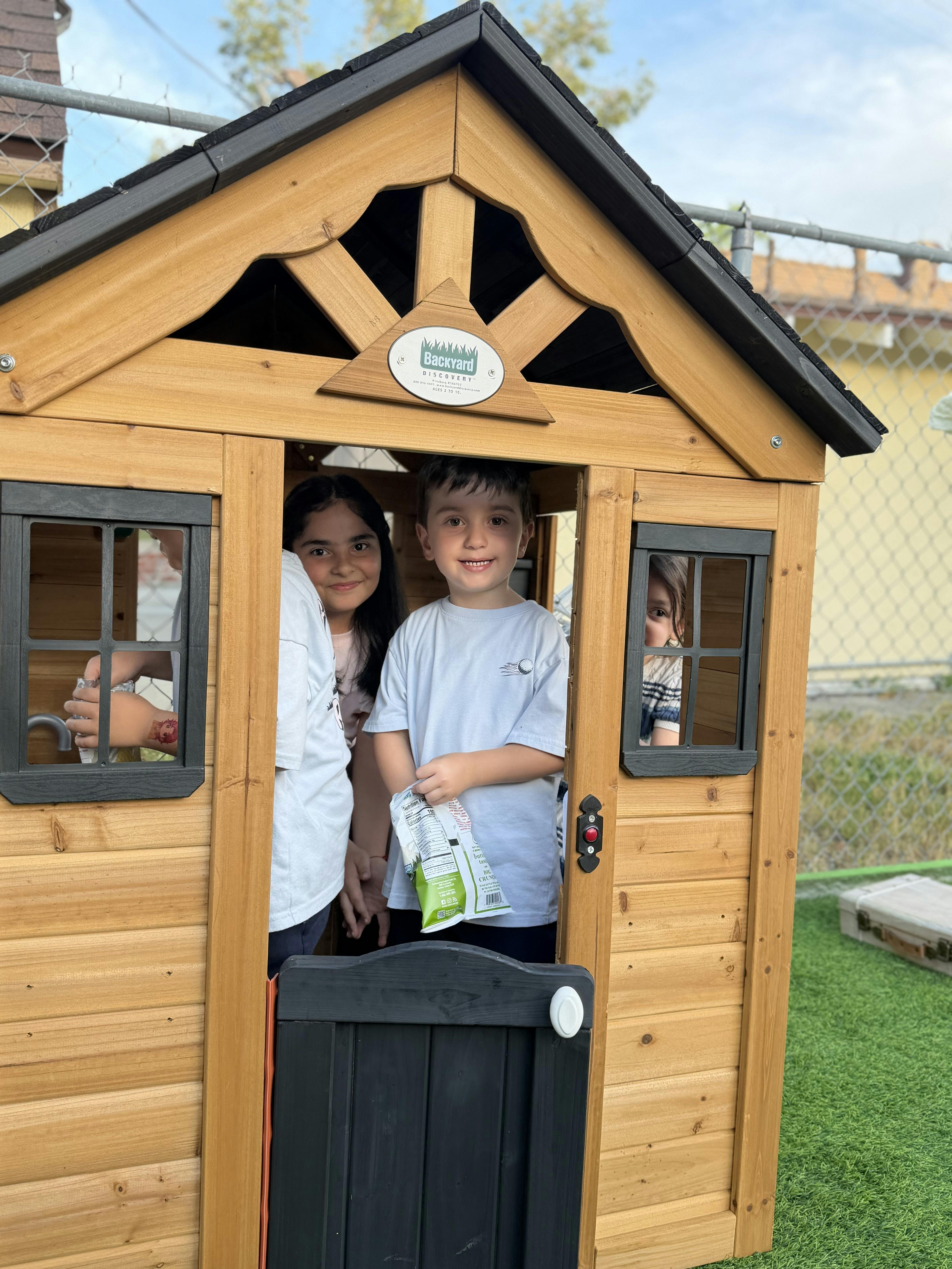 Three children in a wooden playhouse with one holding a snack bag