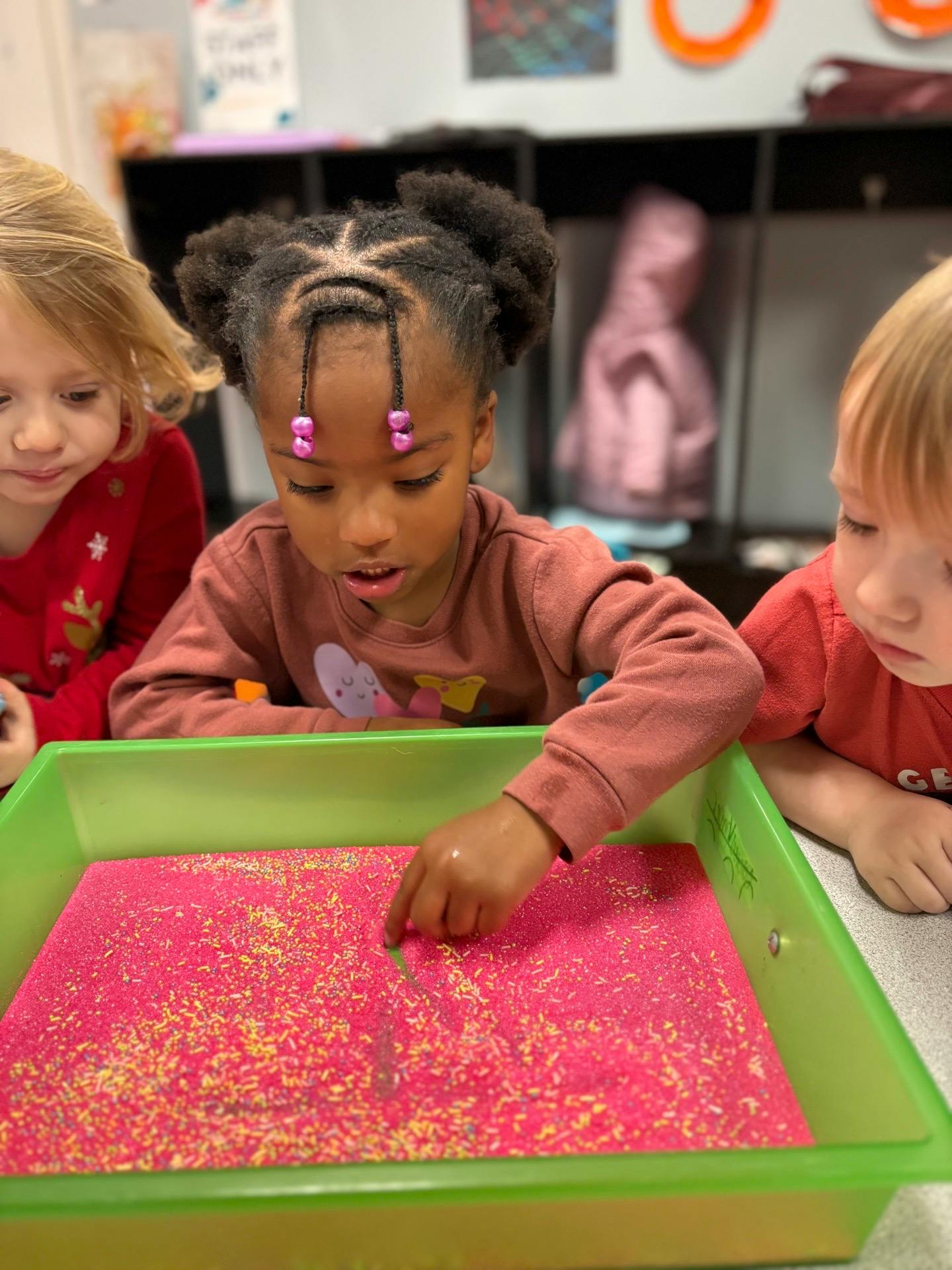 Child drawing in a pink sand tray with two other children