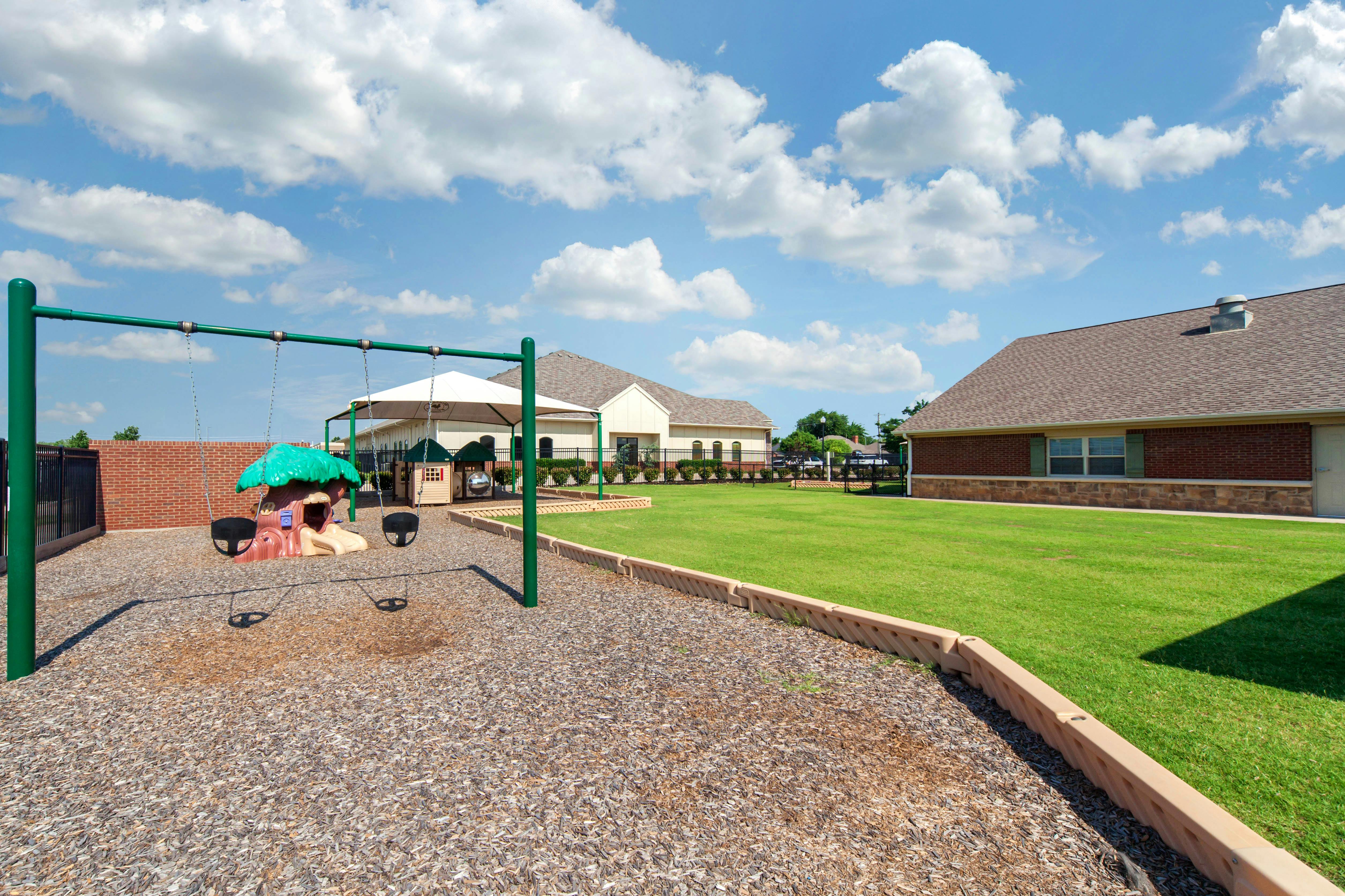 A swing set with a play structure on wood chips next to a grassy area and building