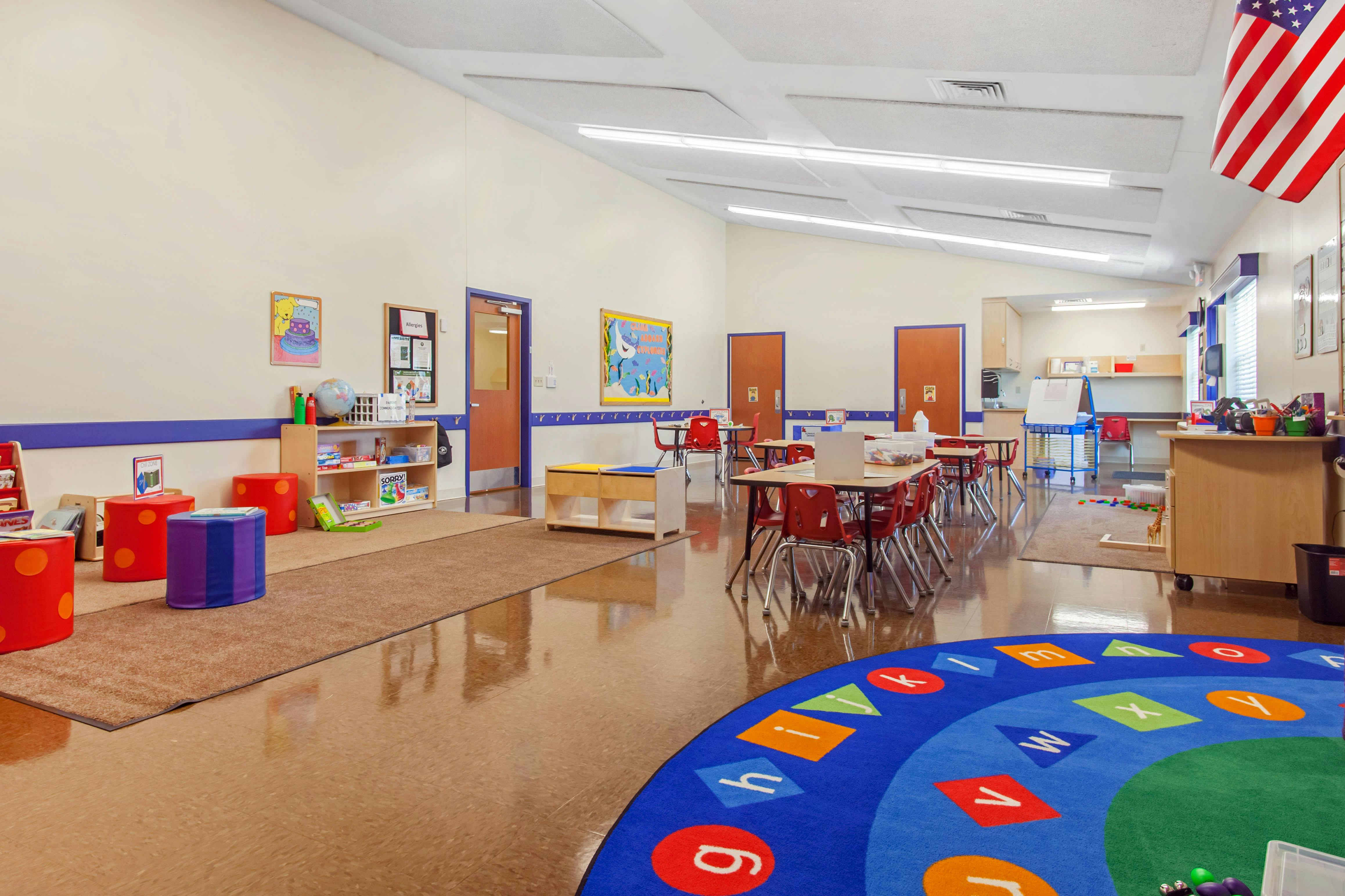 A colorful classroom with small tables and red chairs, a blue alphabet rug