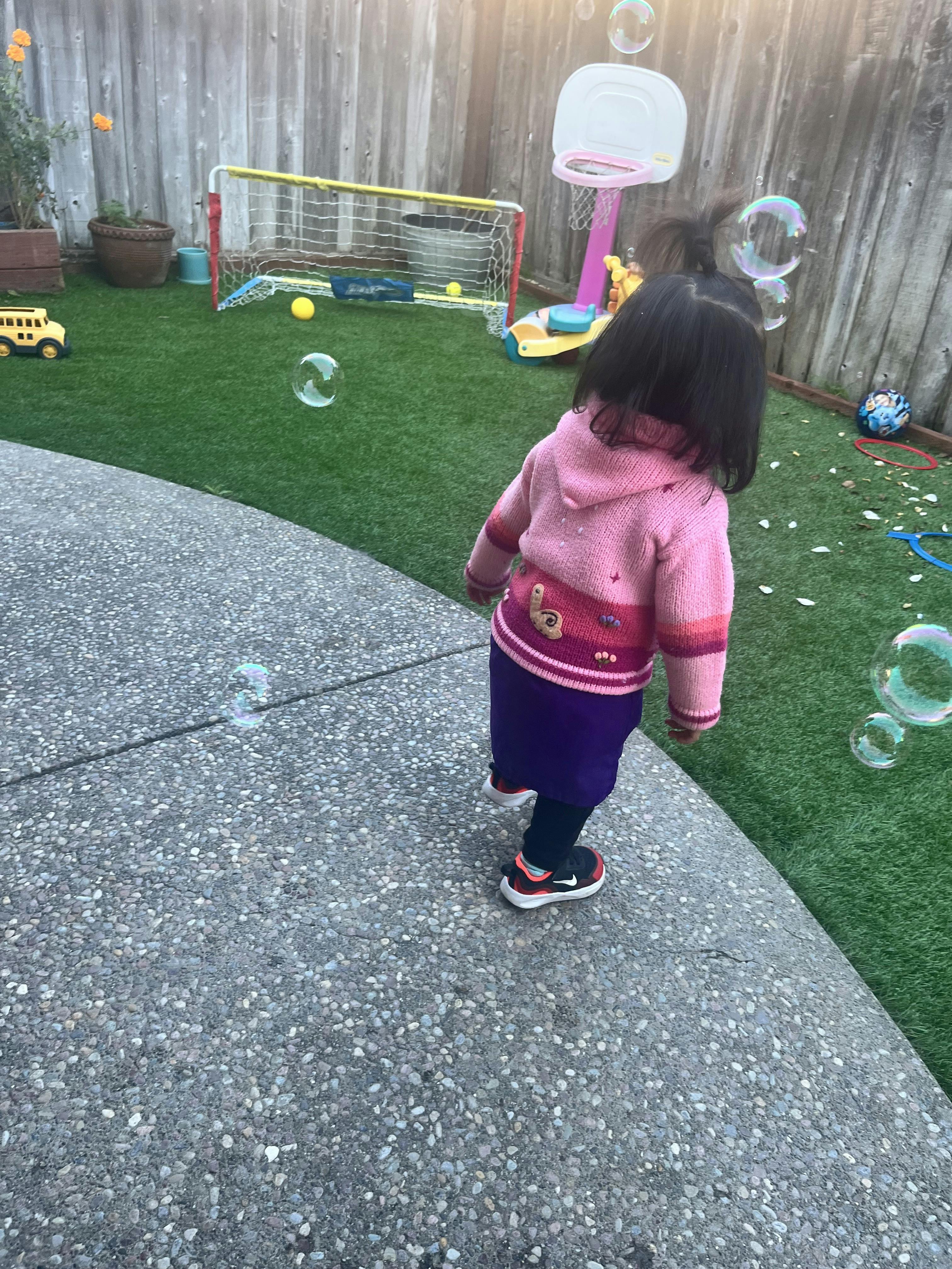 Child in pink sweater watching bubbles in a backyard