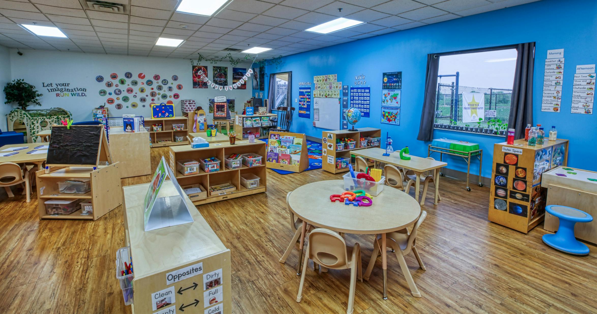 A colorful empty classroom with small tables and chairs, shelves, and educational posters