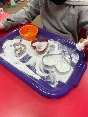 Child playing with heart-shaped cookie cutters and white powder on a tray