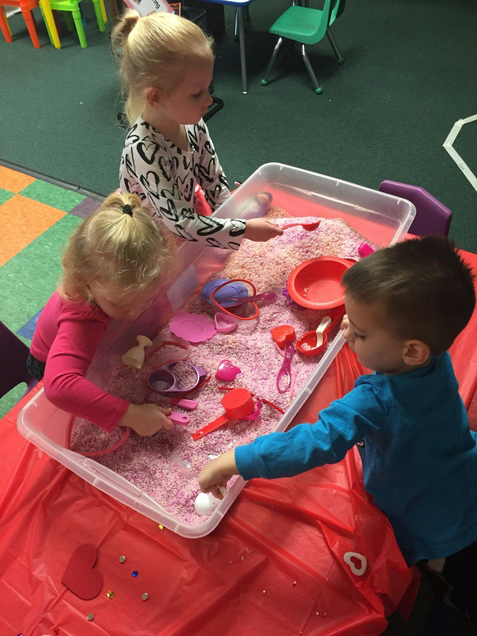 Three children playing with pink rice and toys in a sensory bin