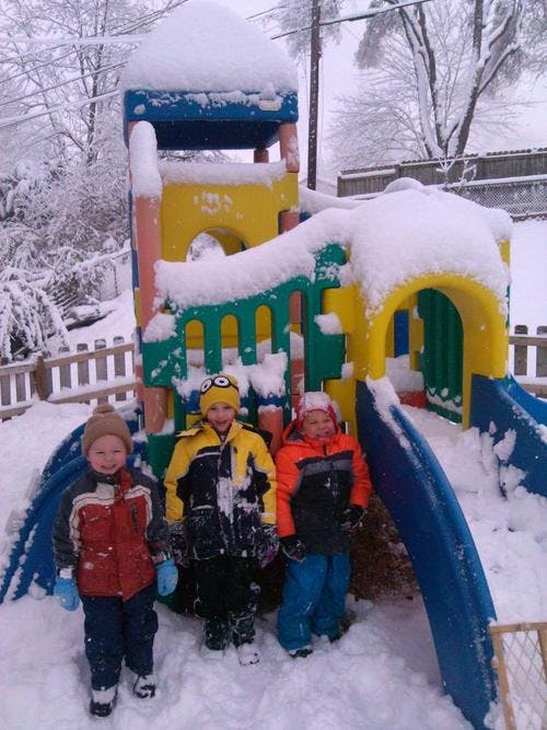 Three children standing in snow next to a snow-covered playground structure
