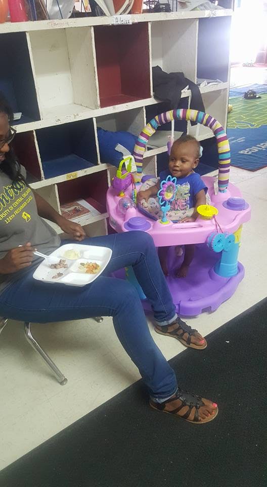 Person eating from a plate on their lap next to a child in a play saucer