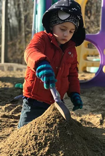 Child in red jacket and gloves digging in sand with a shovel