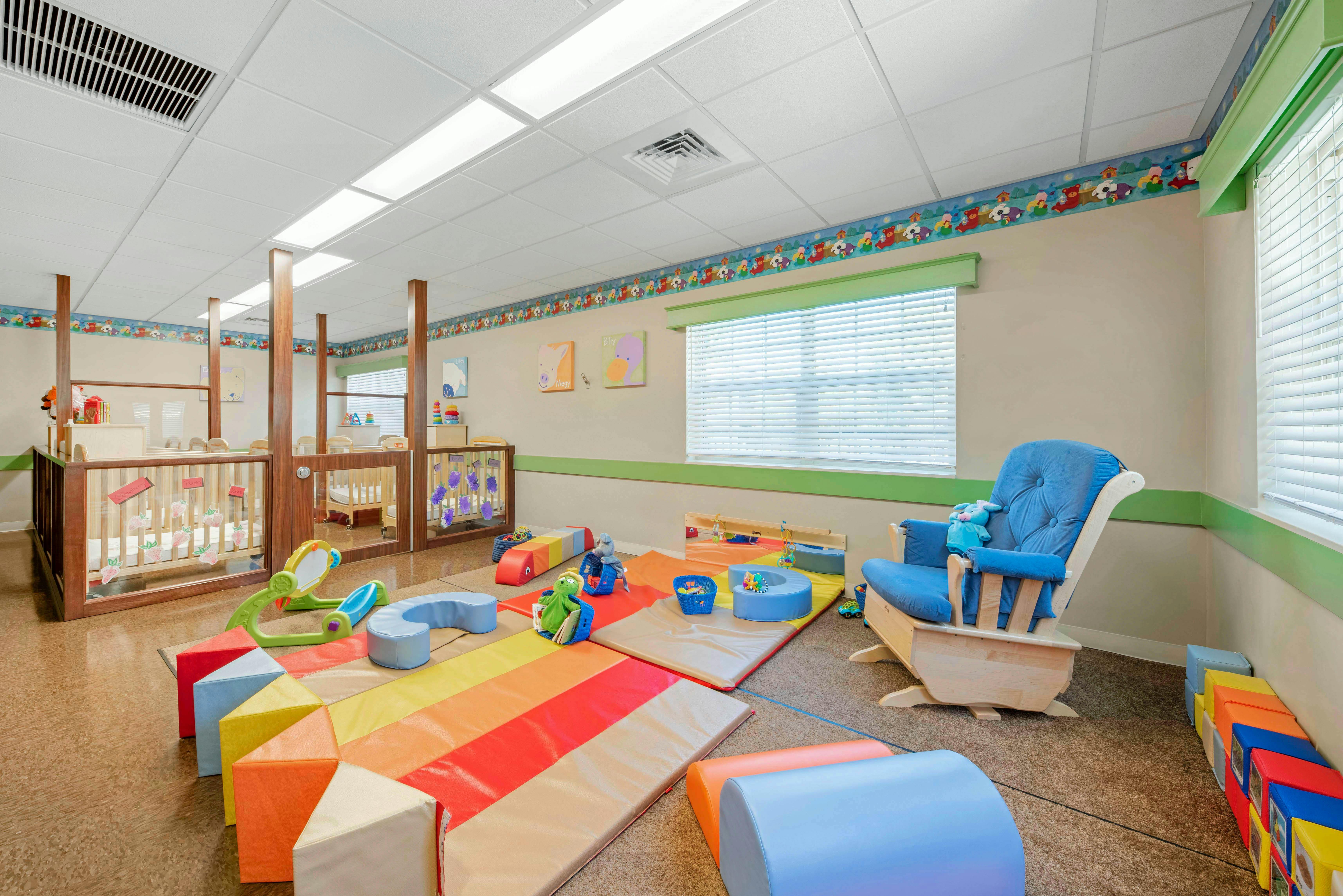 A childcare room with cribs, playmats, soft blocks, and a rocking chair