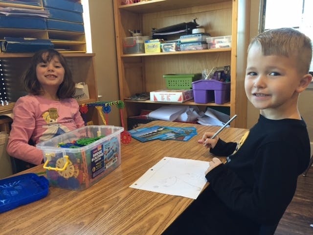Two children at a table, one with a container of toys, one writing