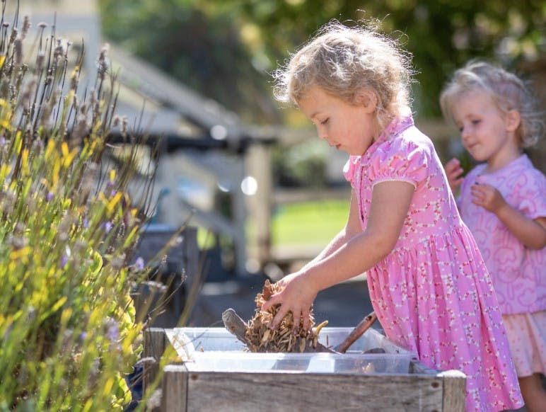 Child playing with wood chips in a container, another child in background