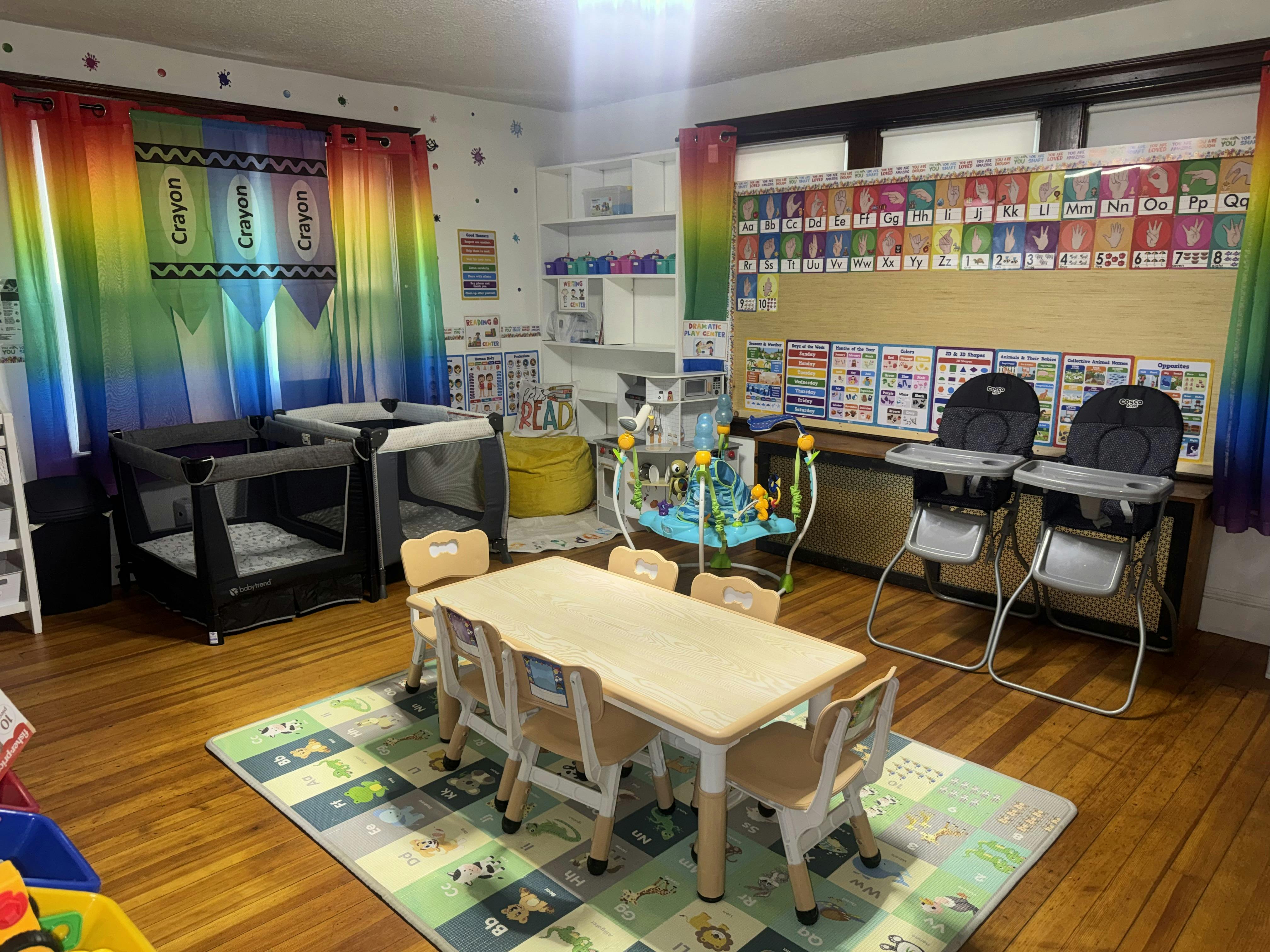 A colorful classroom with playpens, high chairs, a children's table, and learning charts