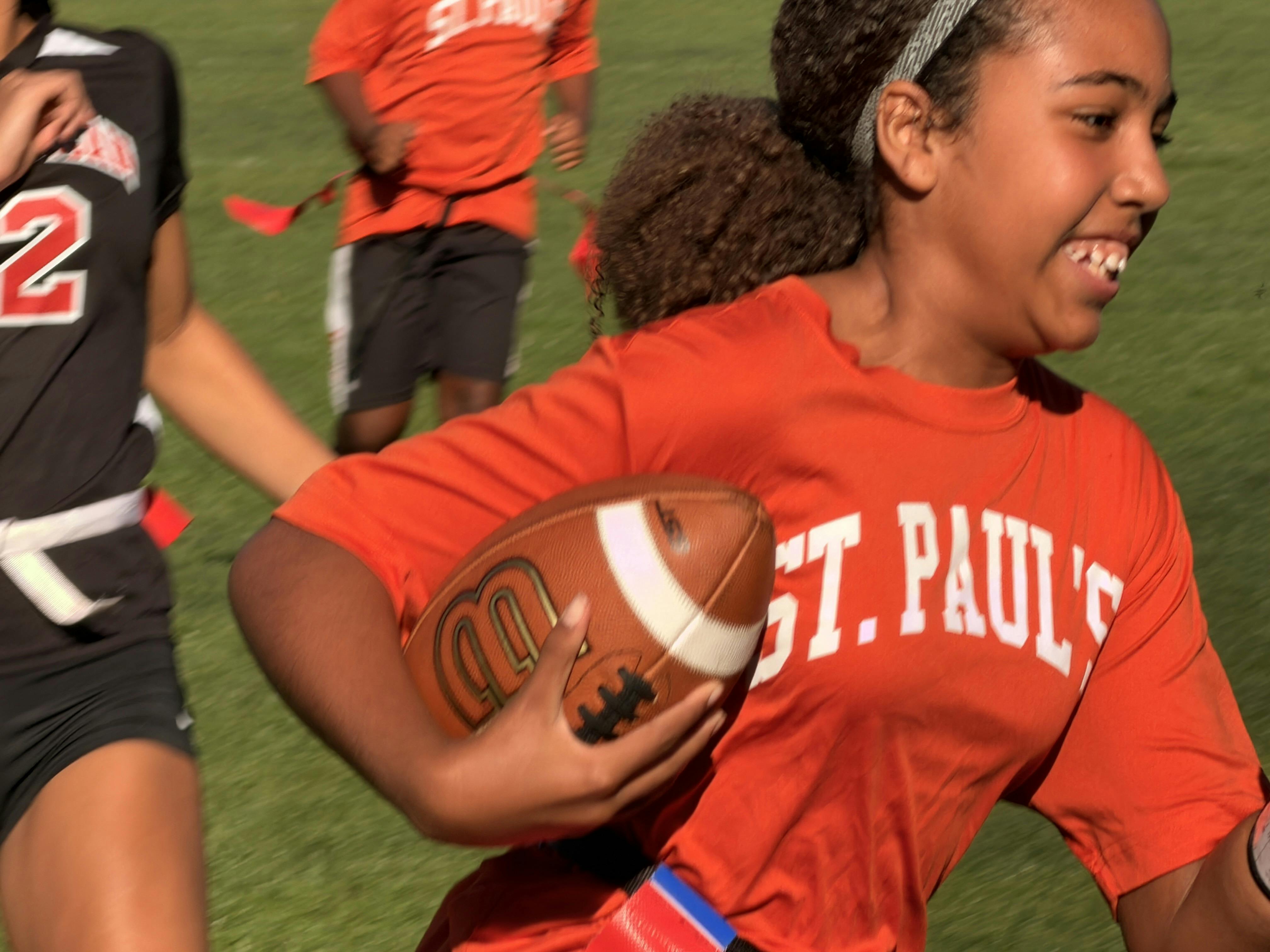 Child in orange jersey with "ST. PAUL'S" running with a football