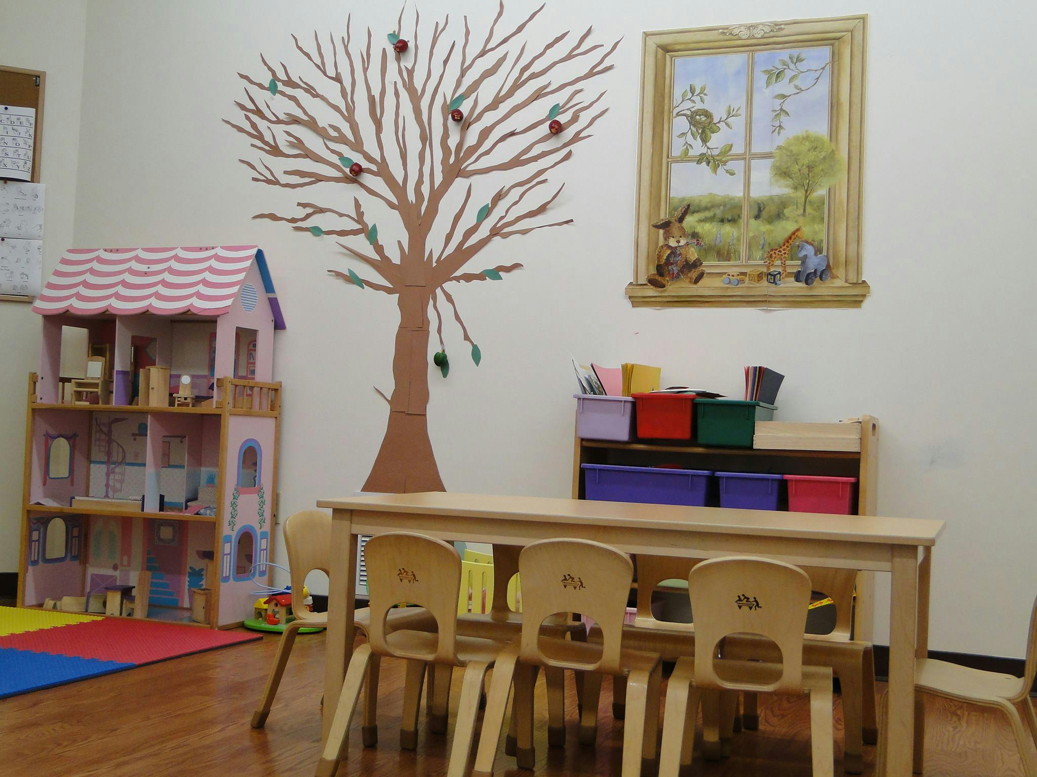 Classroom with a dollhouse, a tree decal on the wall, and a table with chairs