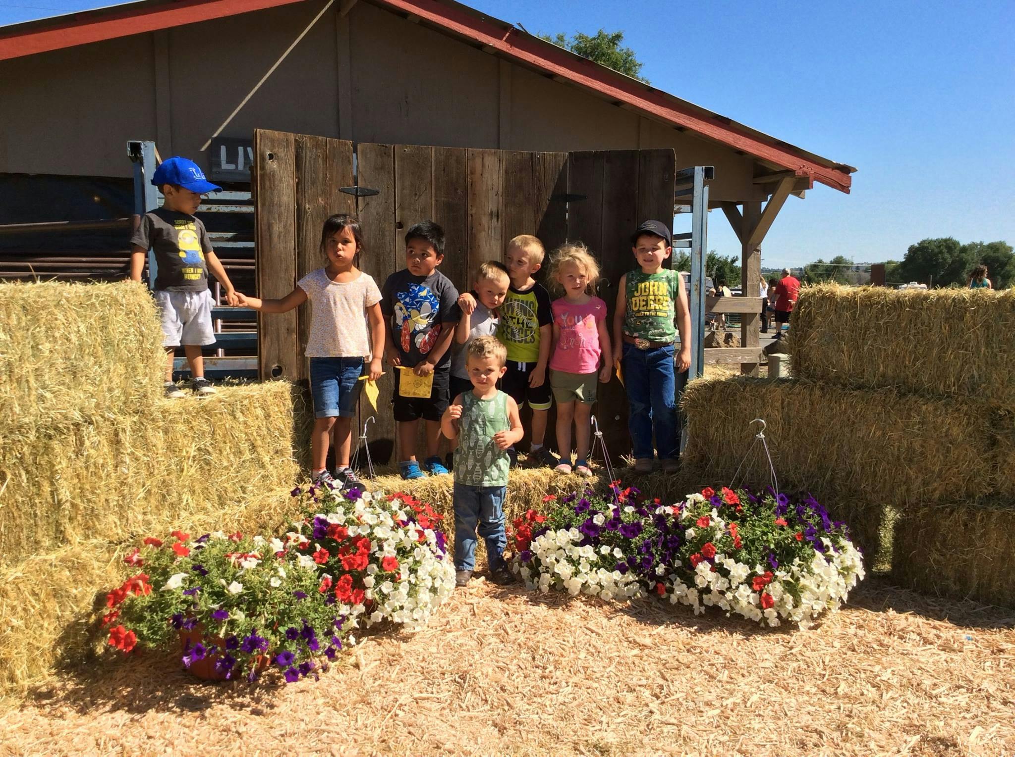 Group of children standing in front of a wooden building with hay bales and flowers