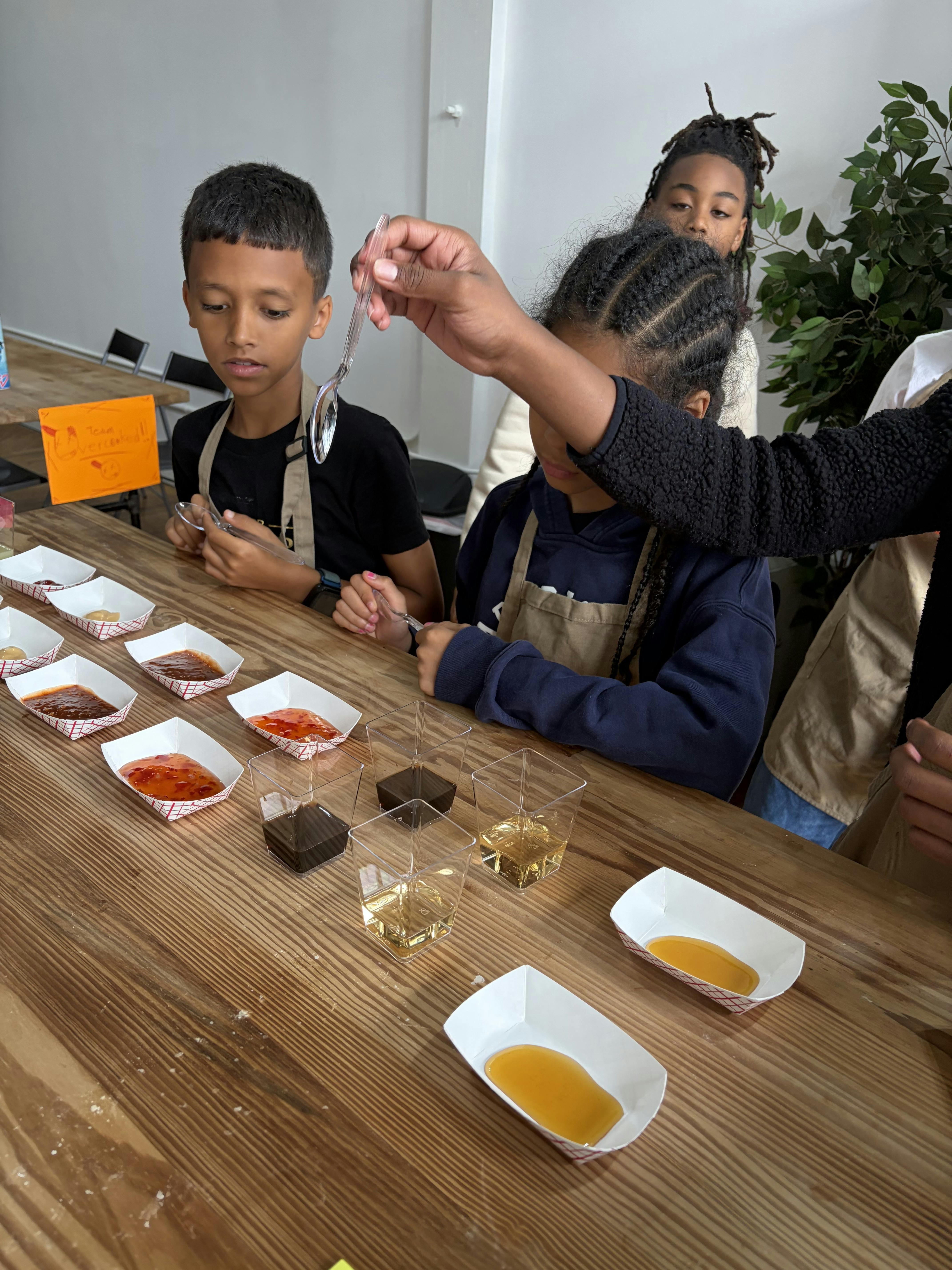 Children tasting various sauces and liquids from small containers at a table