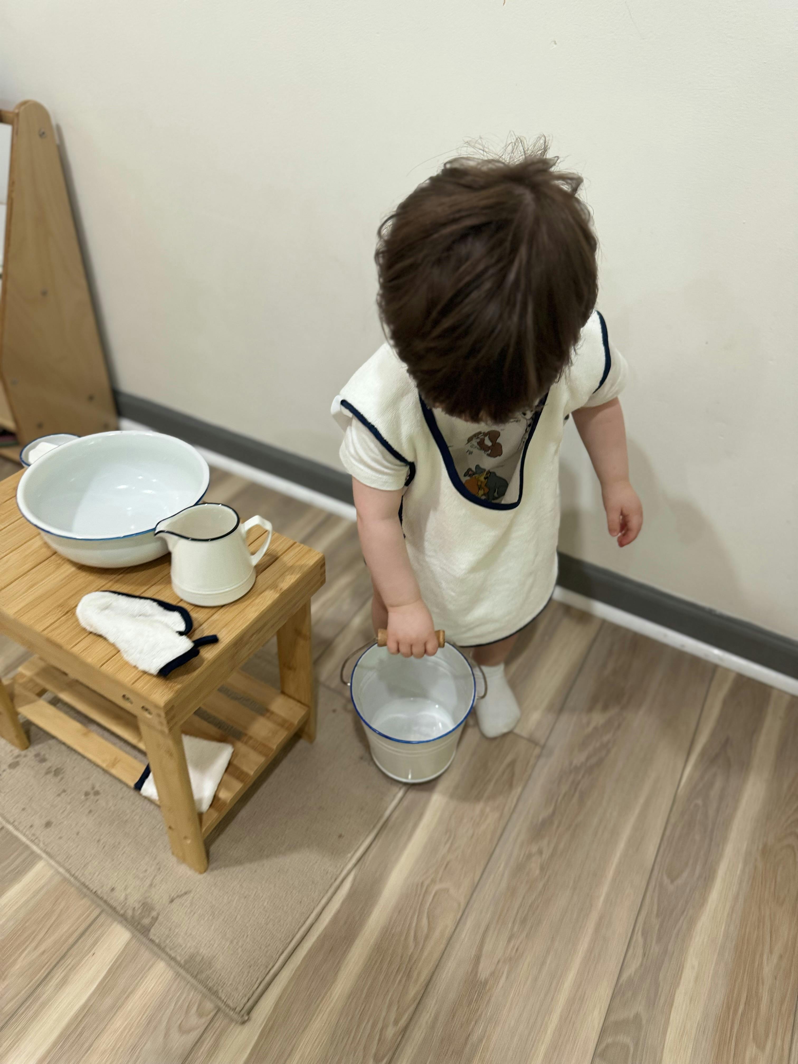 Child holding a small bucket next to a wooden stool with a bowl and pitcher