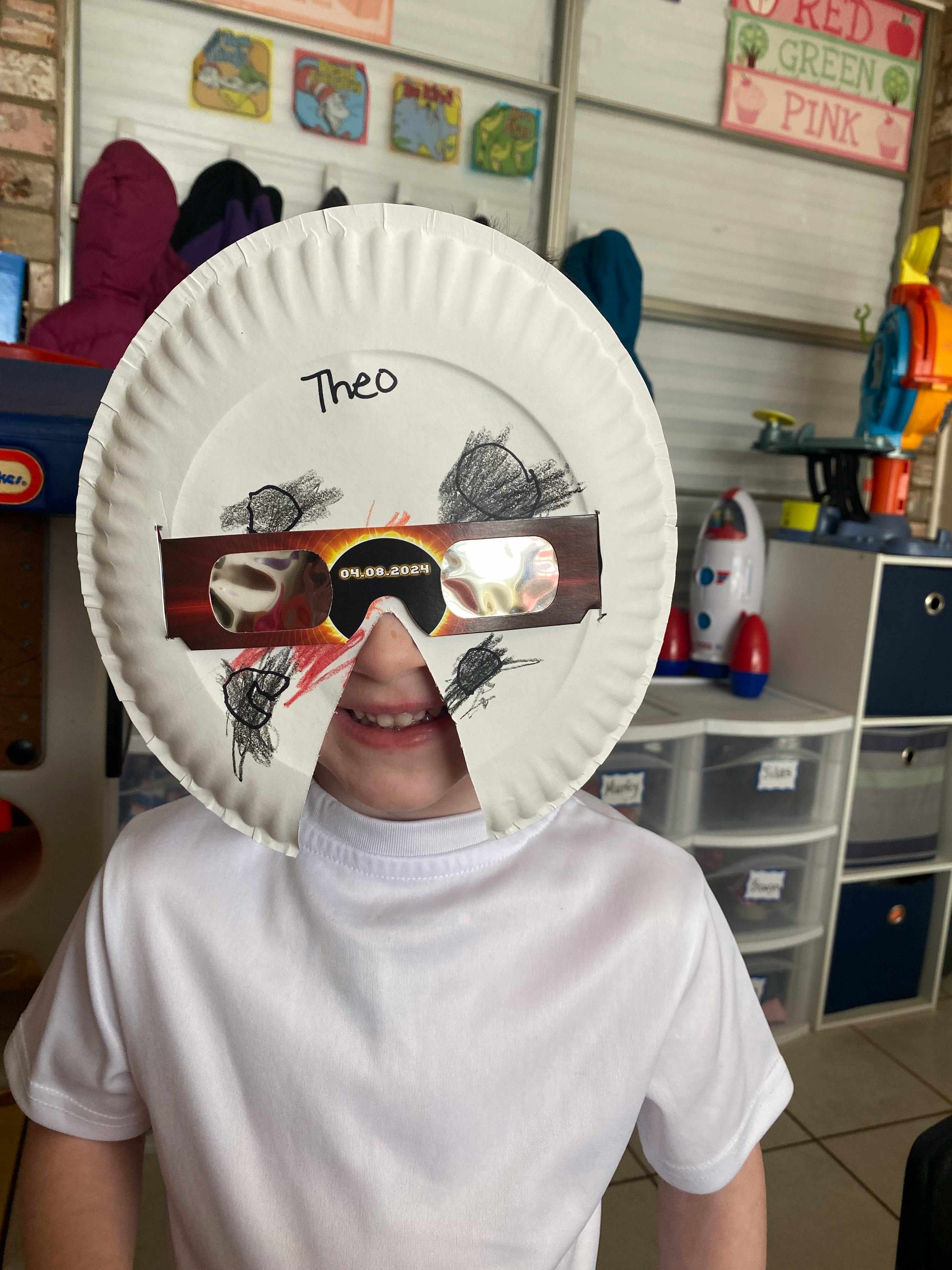 Child wearing a paper plate mask with eclipse glasses and drawings