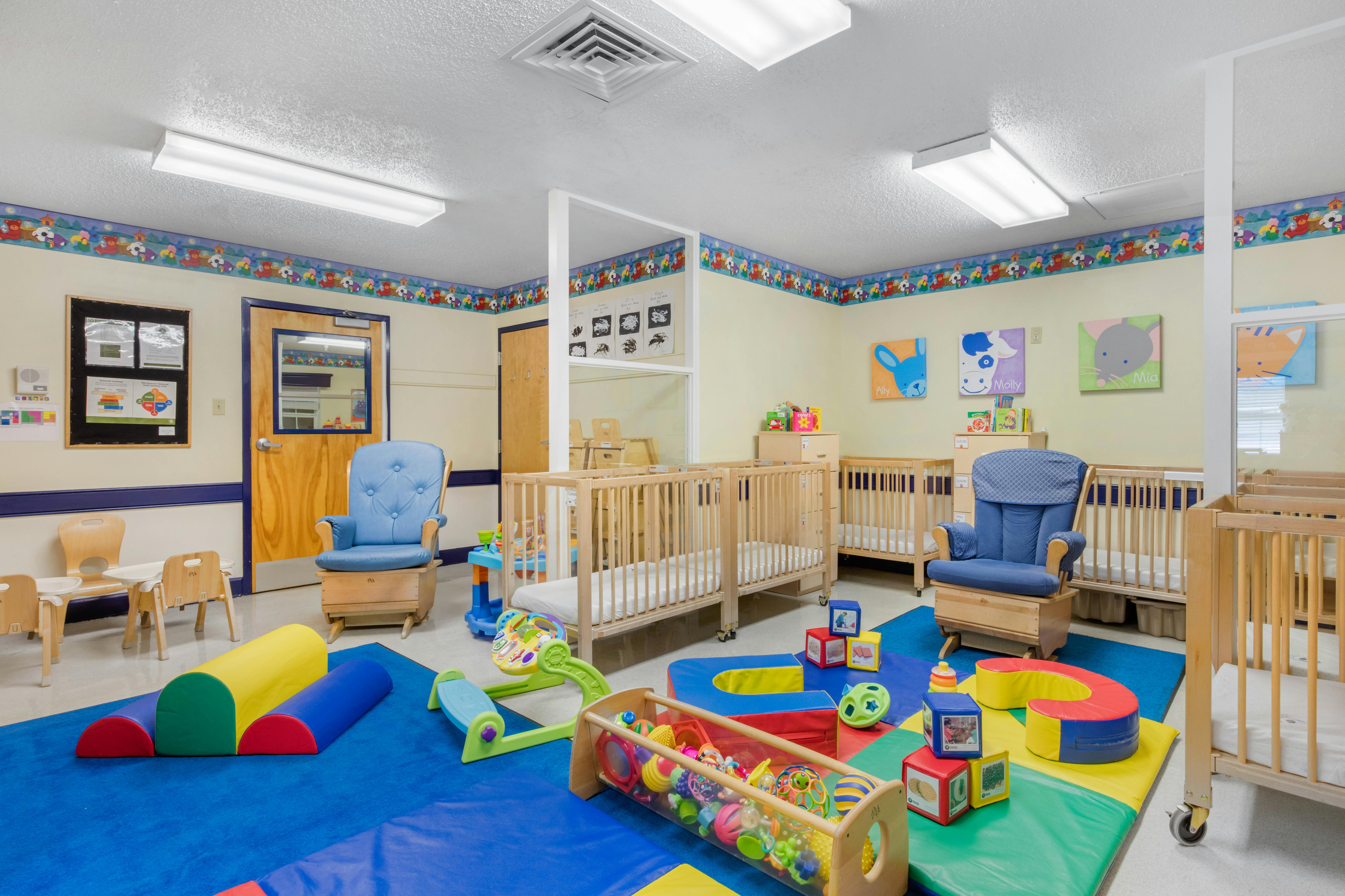 A colorful childcare room with cribs, play mats, and a blue rocking chair