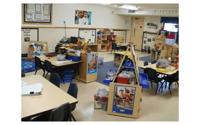 An empty classroom with several tables and chairs, shelves, and learning materials