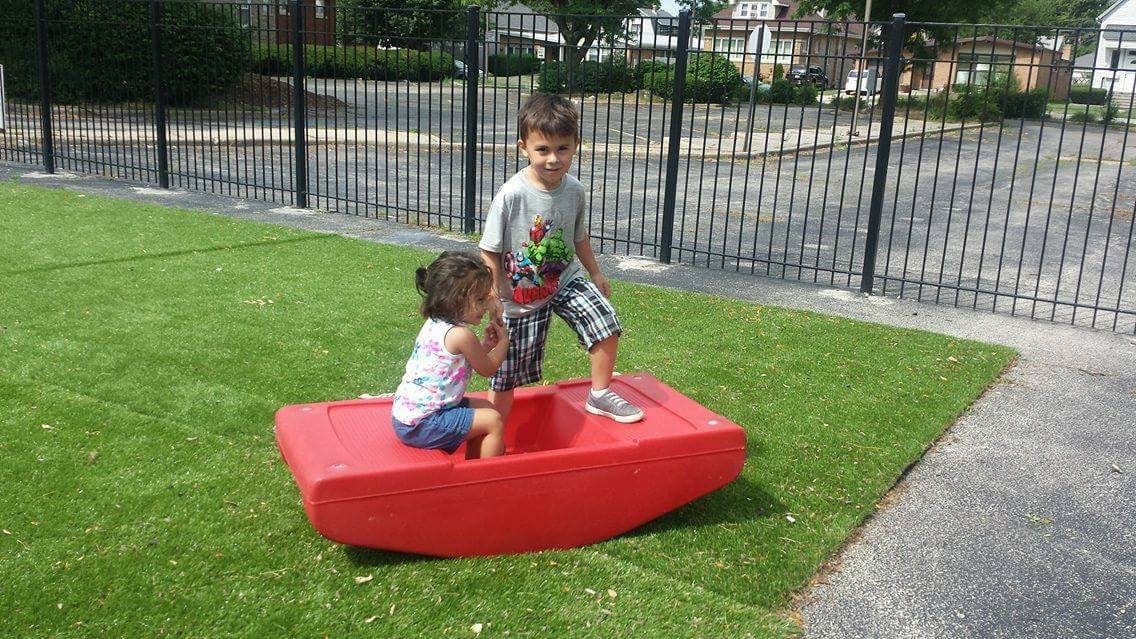 Two children playing on a red boat-shaped toy on grass