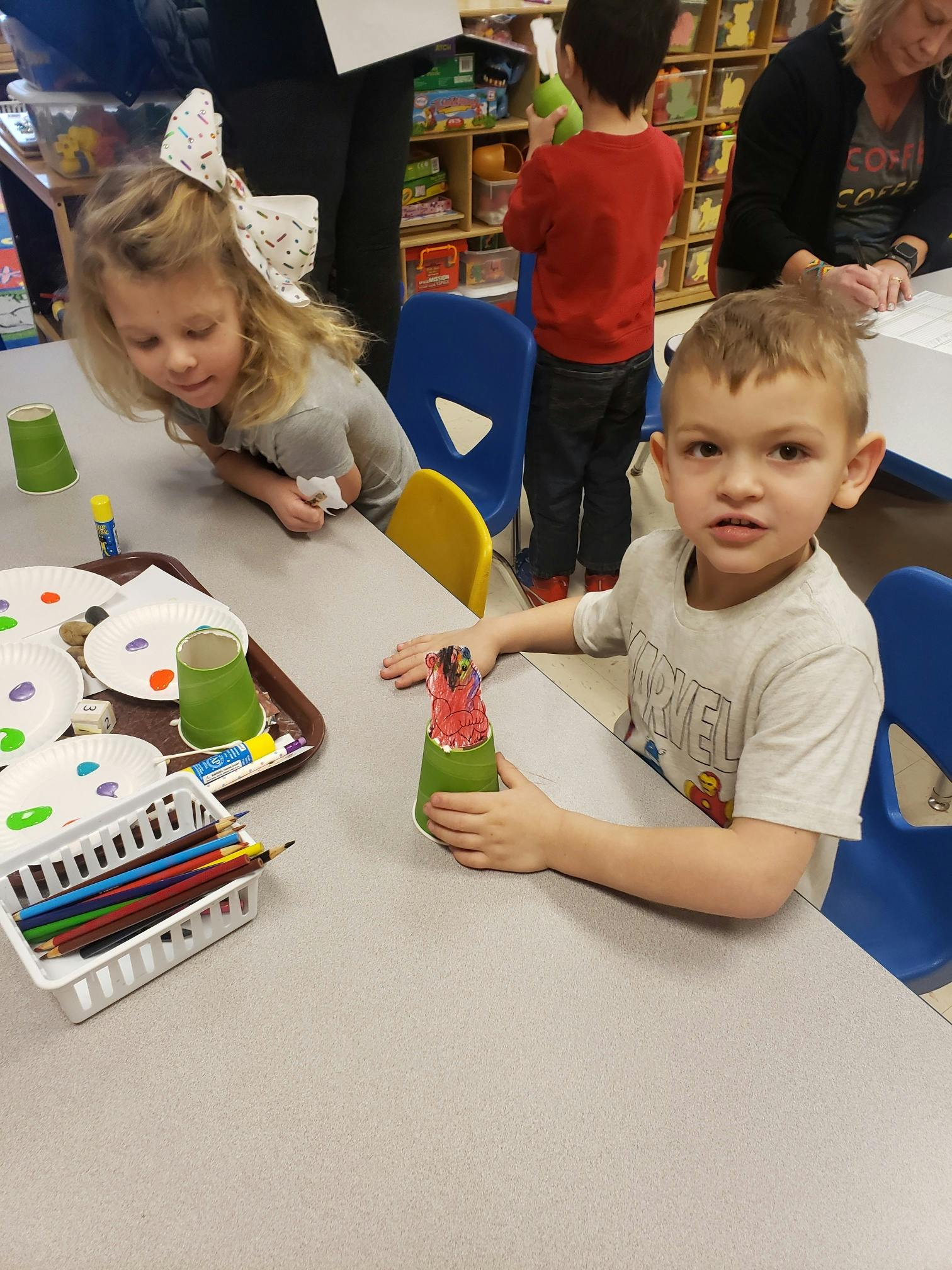 Child holding a craft at a table with another child looking on the background