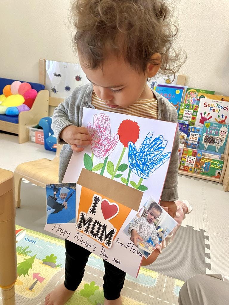 Child holding a handmade card with handprints and "I love Mom" text