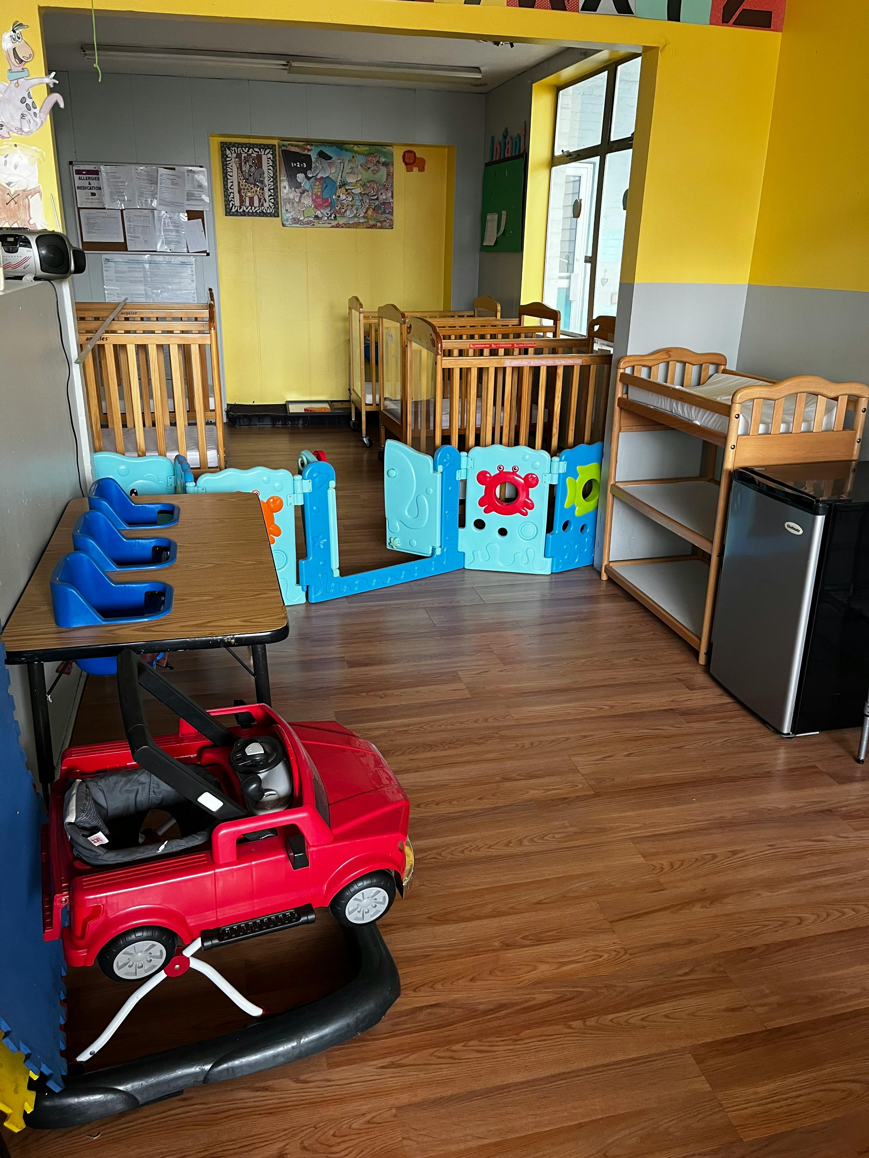Daycare room with cribs, playpen, table with blue seats, and red toy car