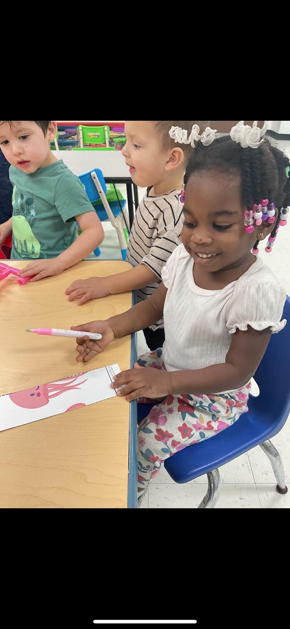 Child holding a pink marker at a table with other children