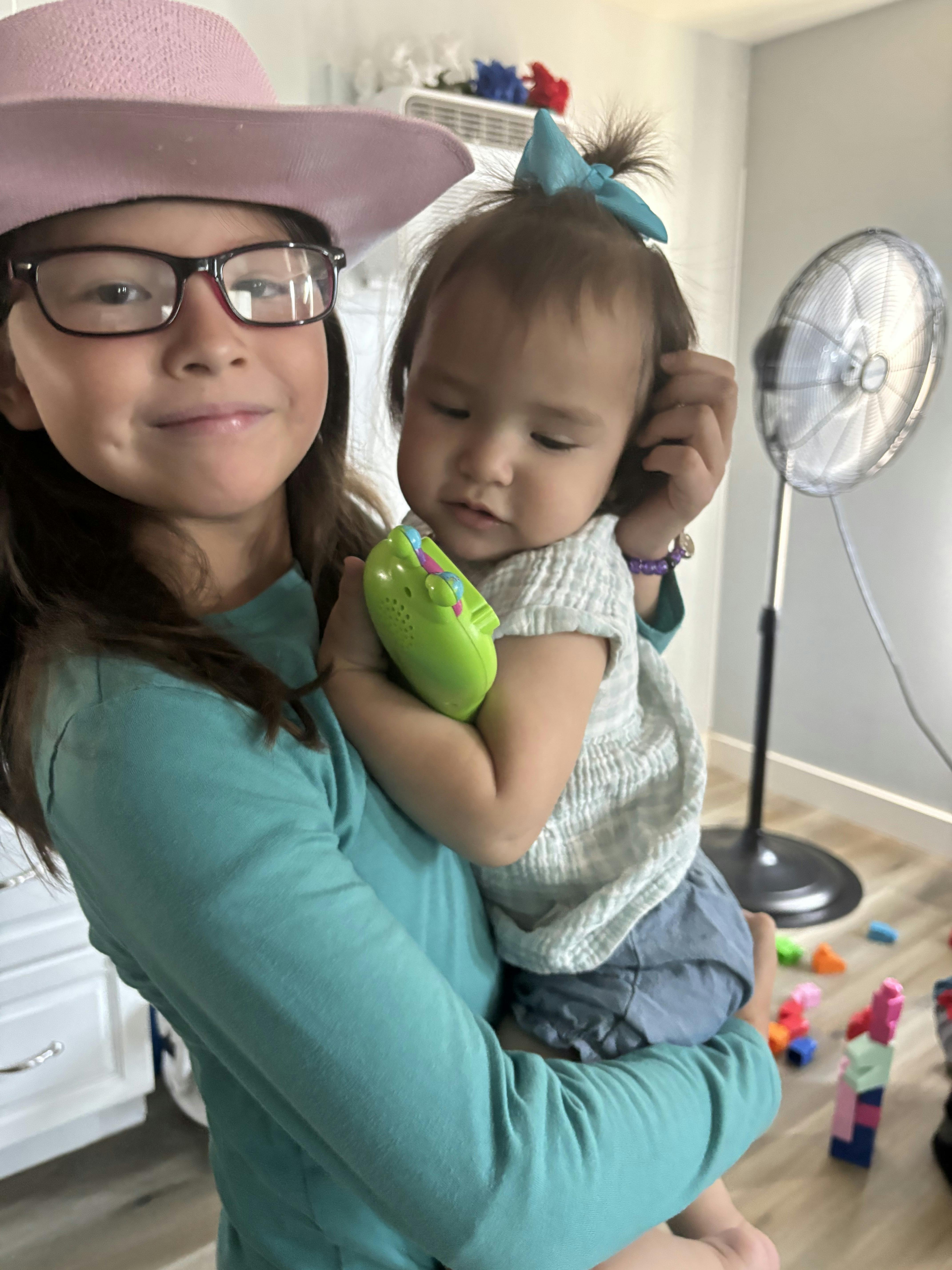 Child in glasses and hat holding a baby with a toy