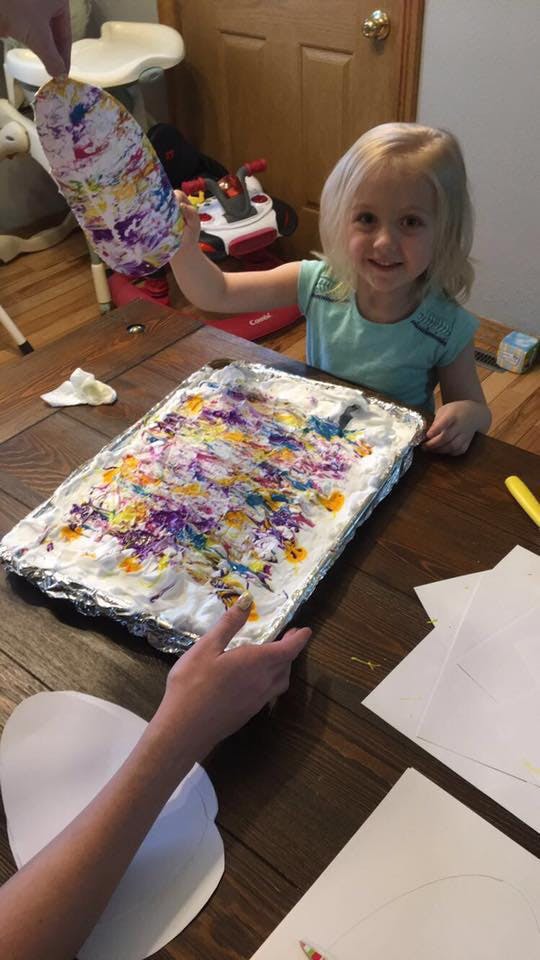 Child holding up a colorful painted paper cutout, person holding a tray of paint