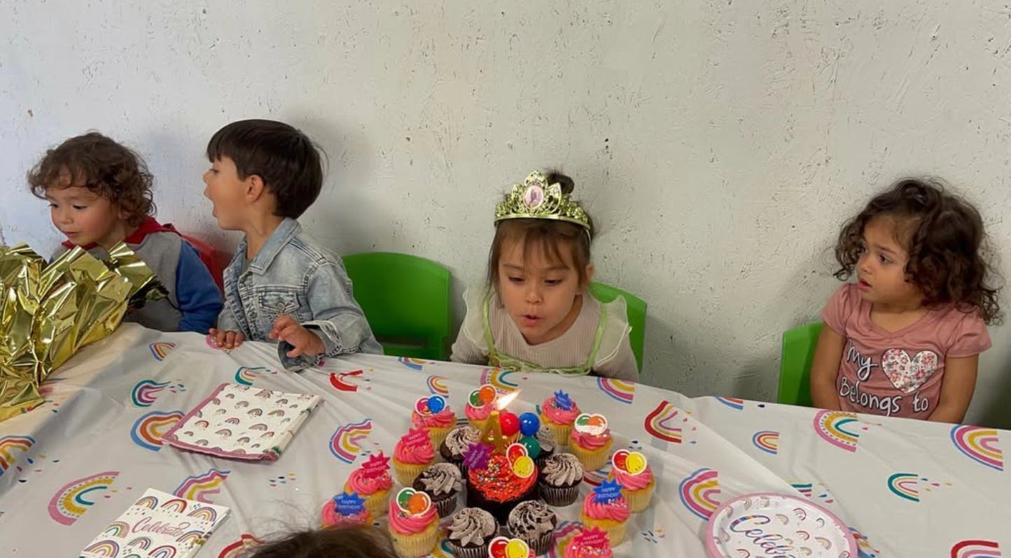 Child in crown blowing out candles on cupcakes at a table with other children