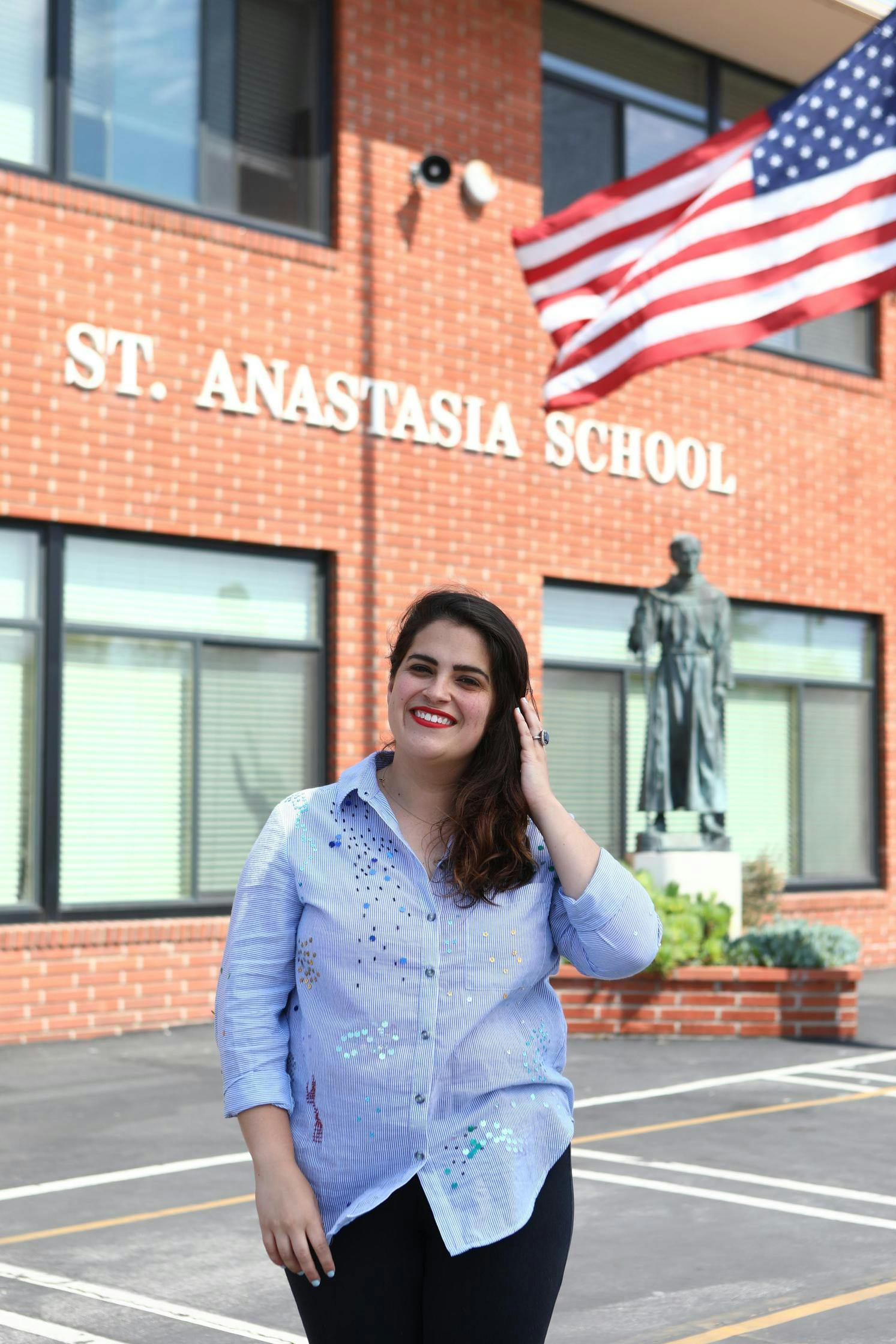 Person in a striped shirt standing in front of a brick school building with an American flag