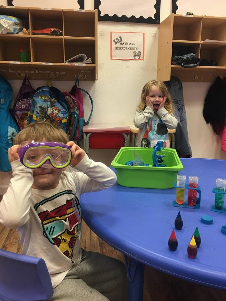 Child holding goggles, another child behind a table with science experiment items