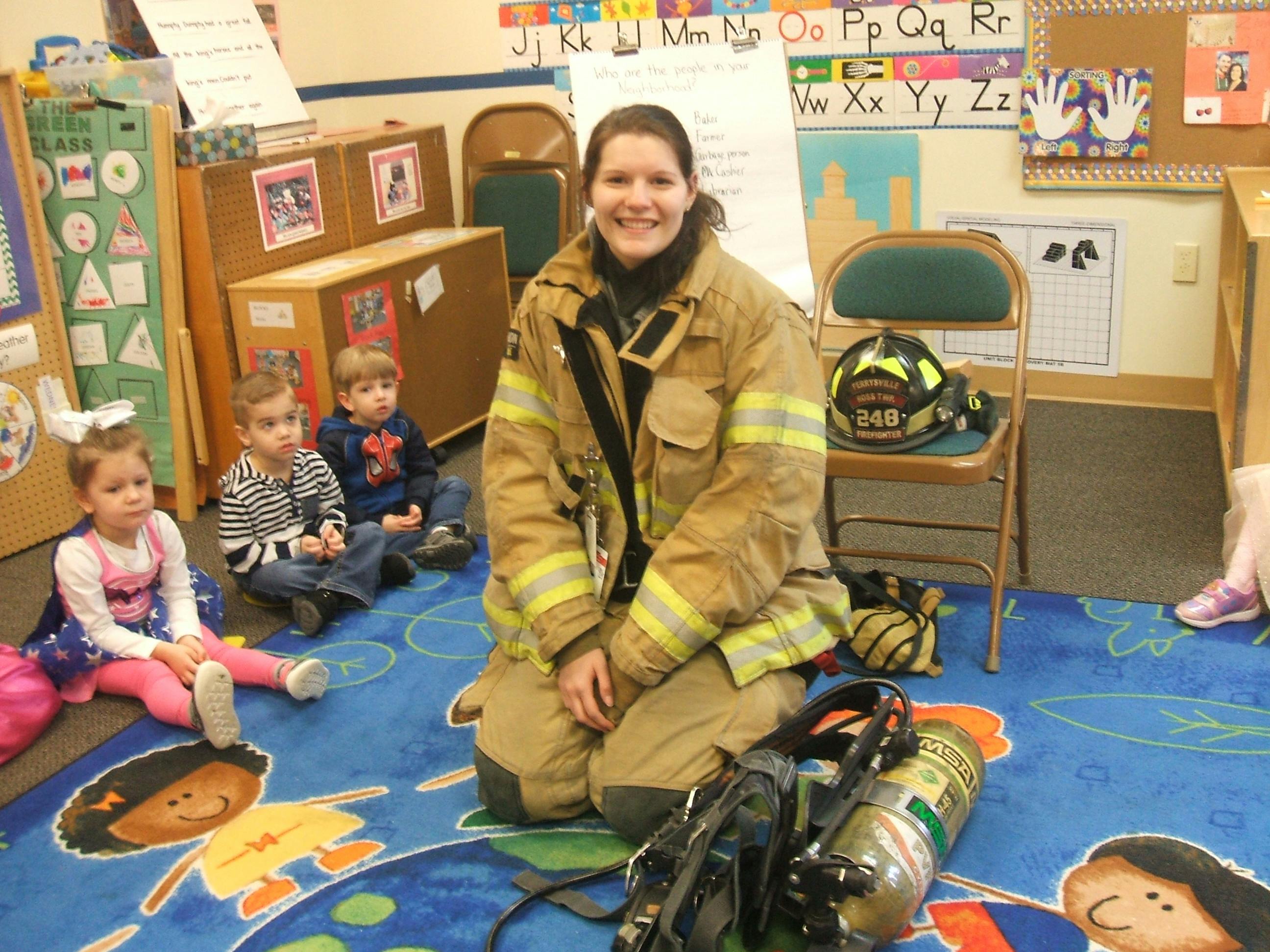 Person in firefighter gear kneeling with three children in a classroom