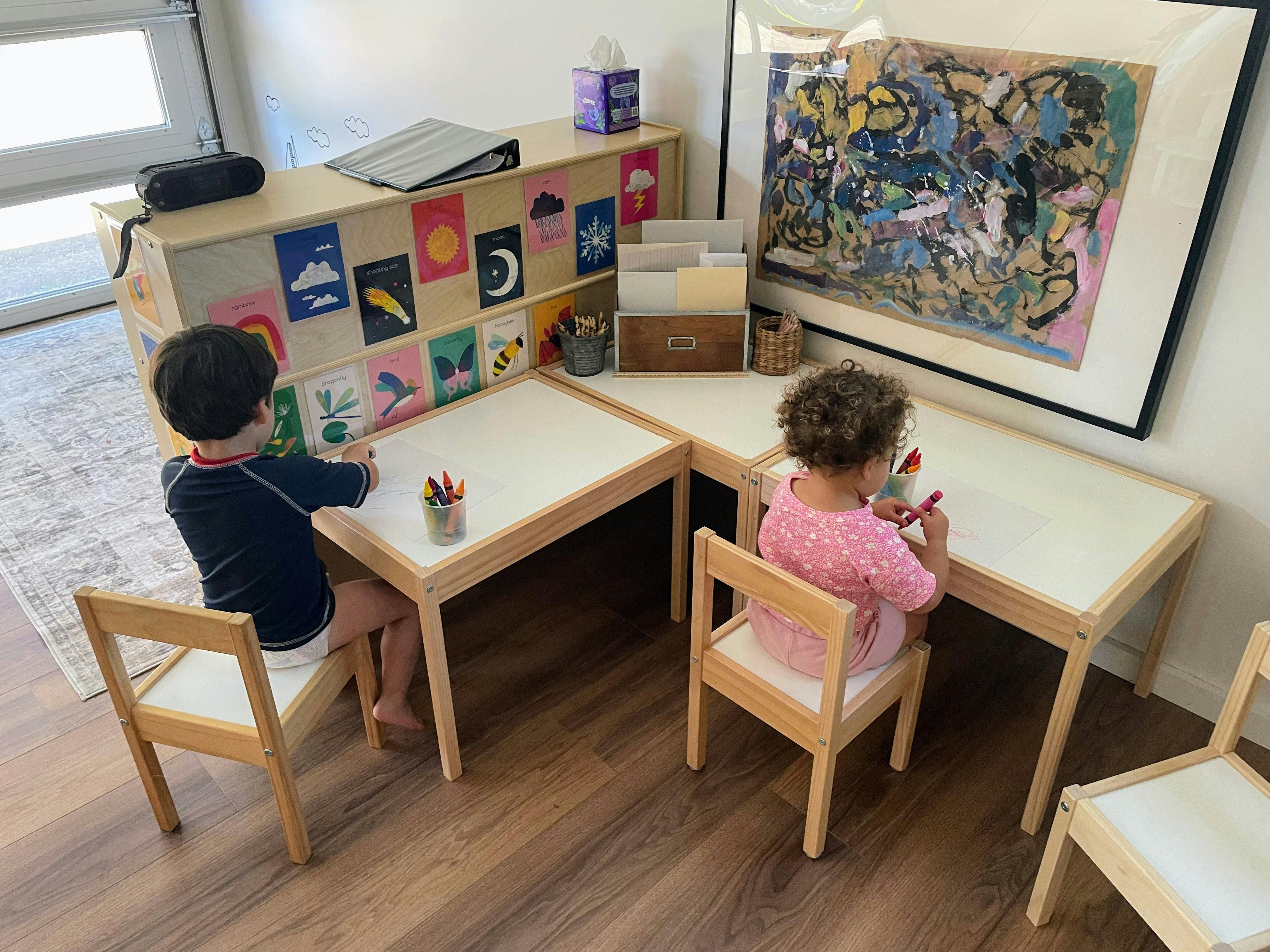 Two children sitting at small tables drawing with crayons in a room