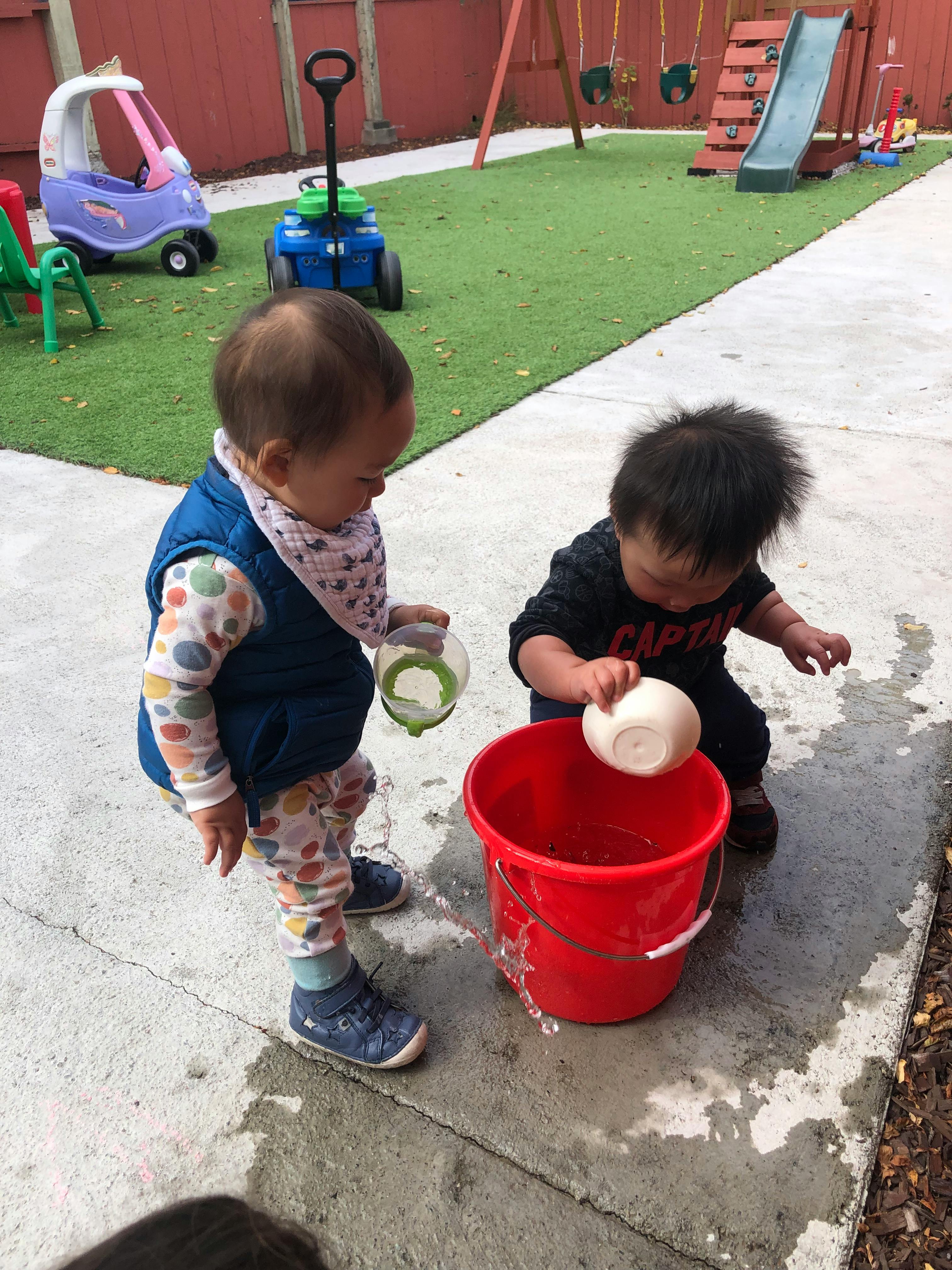 Two children playing with water and a red bucket on a patio