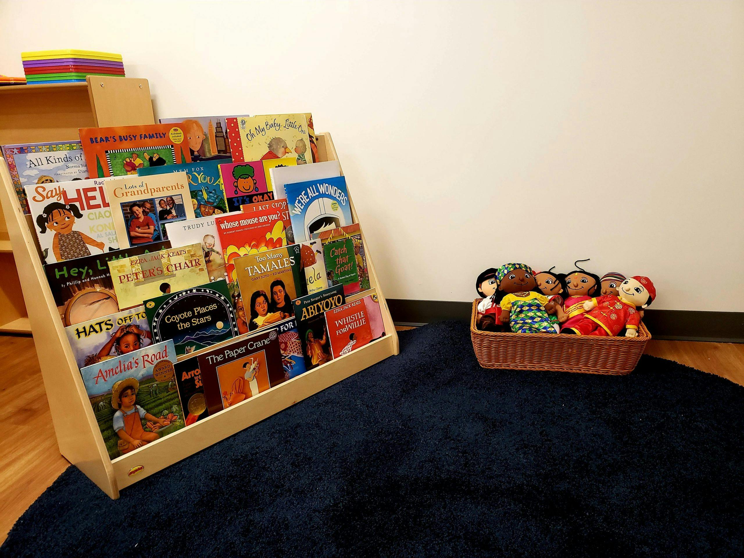 Book display with children's books and a basket of dolls on a rug