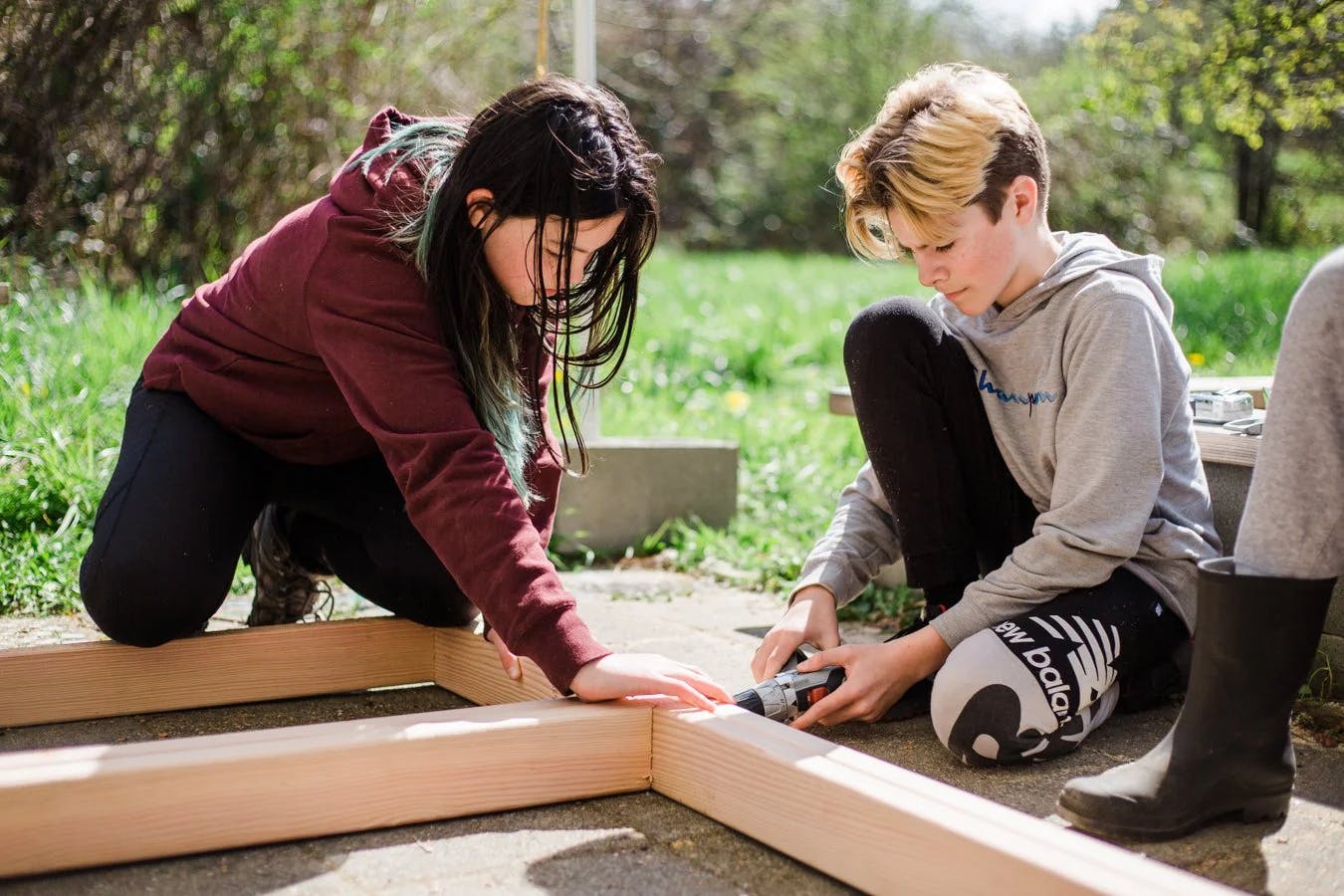 Two teens assembling a wooden frame outdoors, one holding a piece, the other using a drill