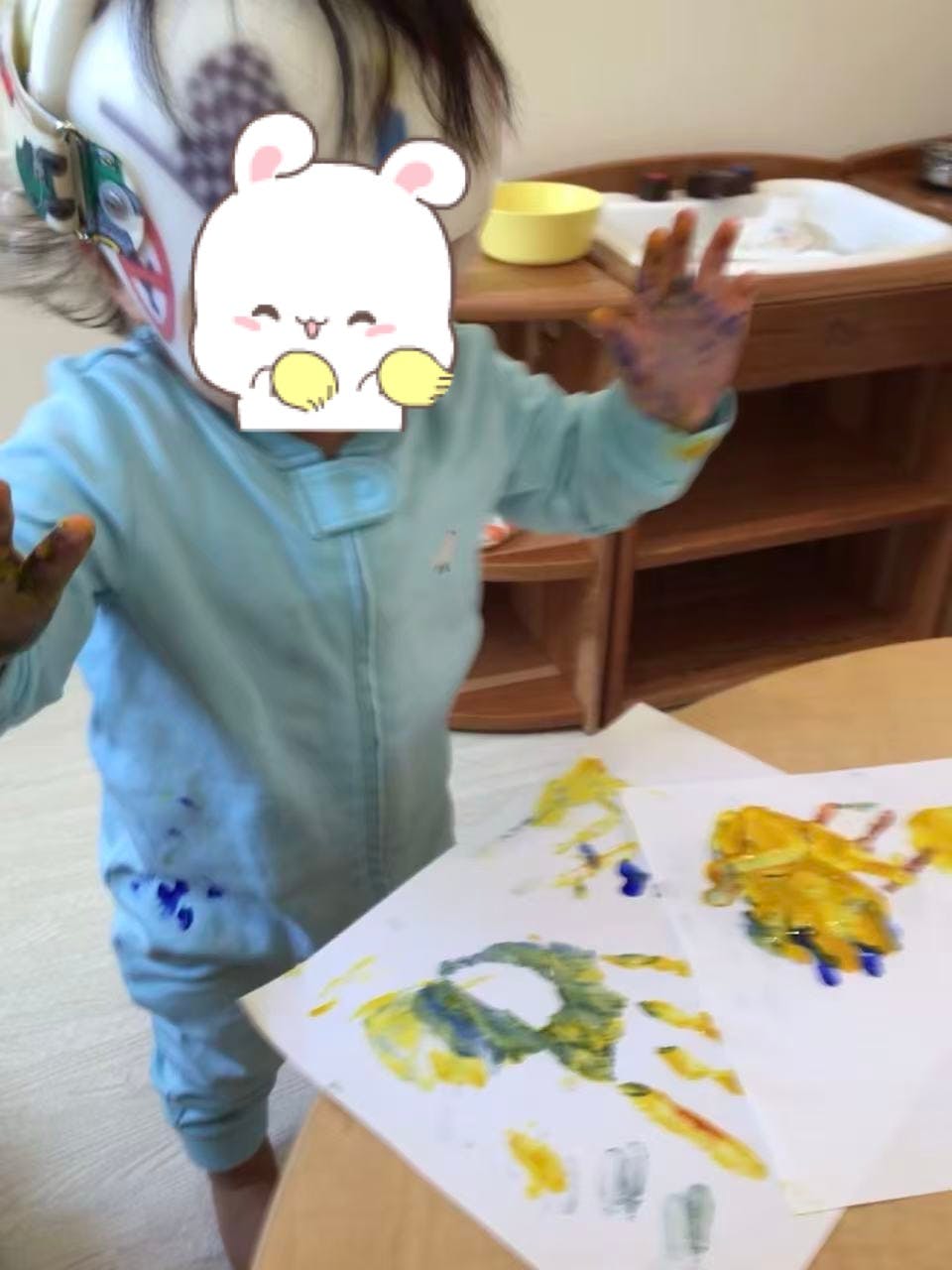 Child with paint on hands and clothes next to paper with painted handprints
