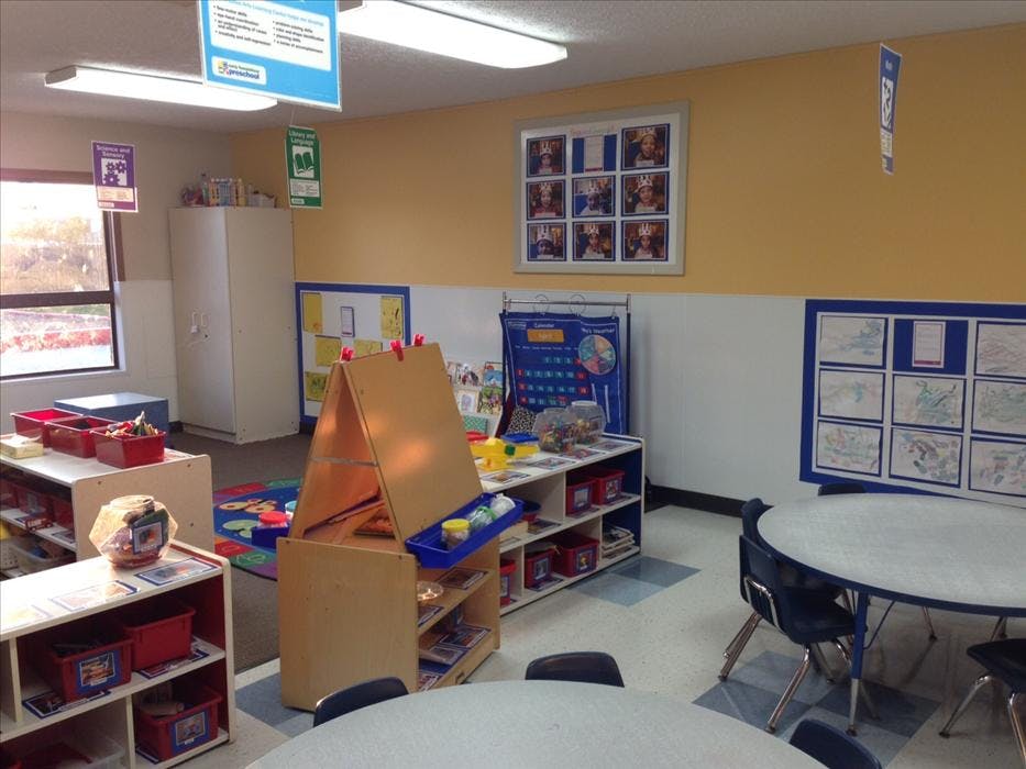 An empty classroom with small tables and chairs, shelves, and a calendar