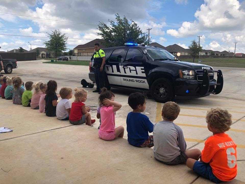 Children sitting in a line watching a person next to a police vehicle