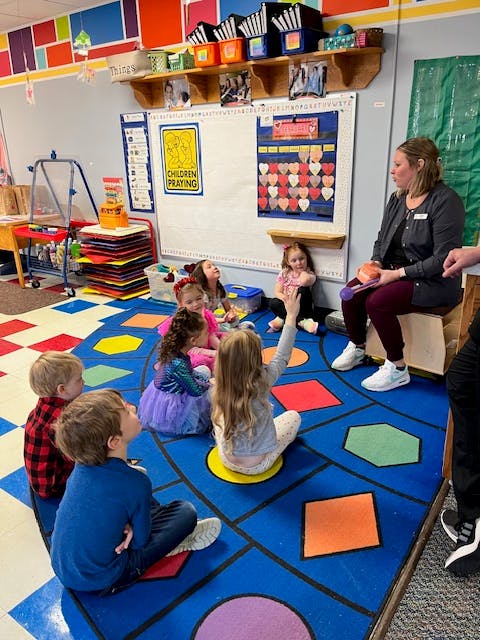Adult speaking to children sitting on a colorful rug in a classroom