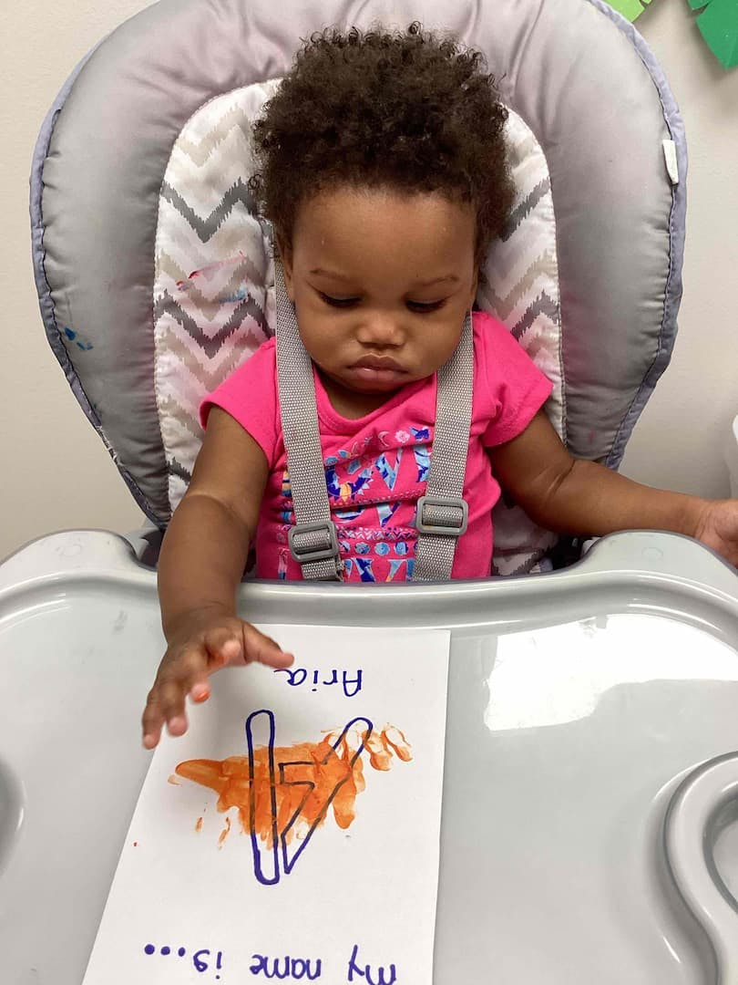 Child in a high chair reaching for a paper with text and an orange handprint