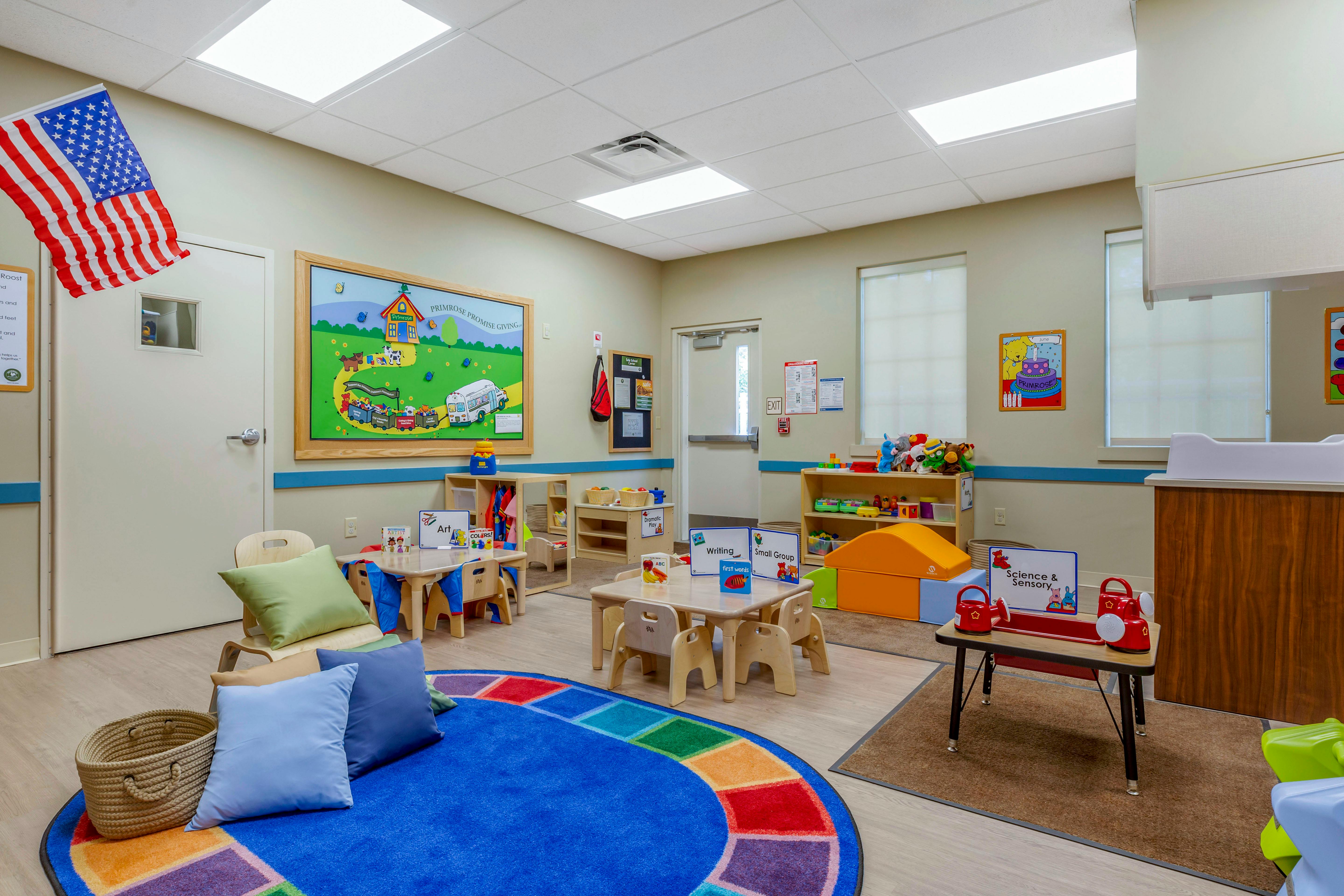 An empty classroom with small wooden tables and chairs, a colorful rug, and toys