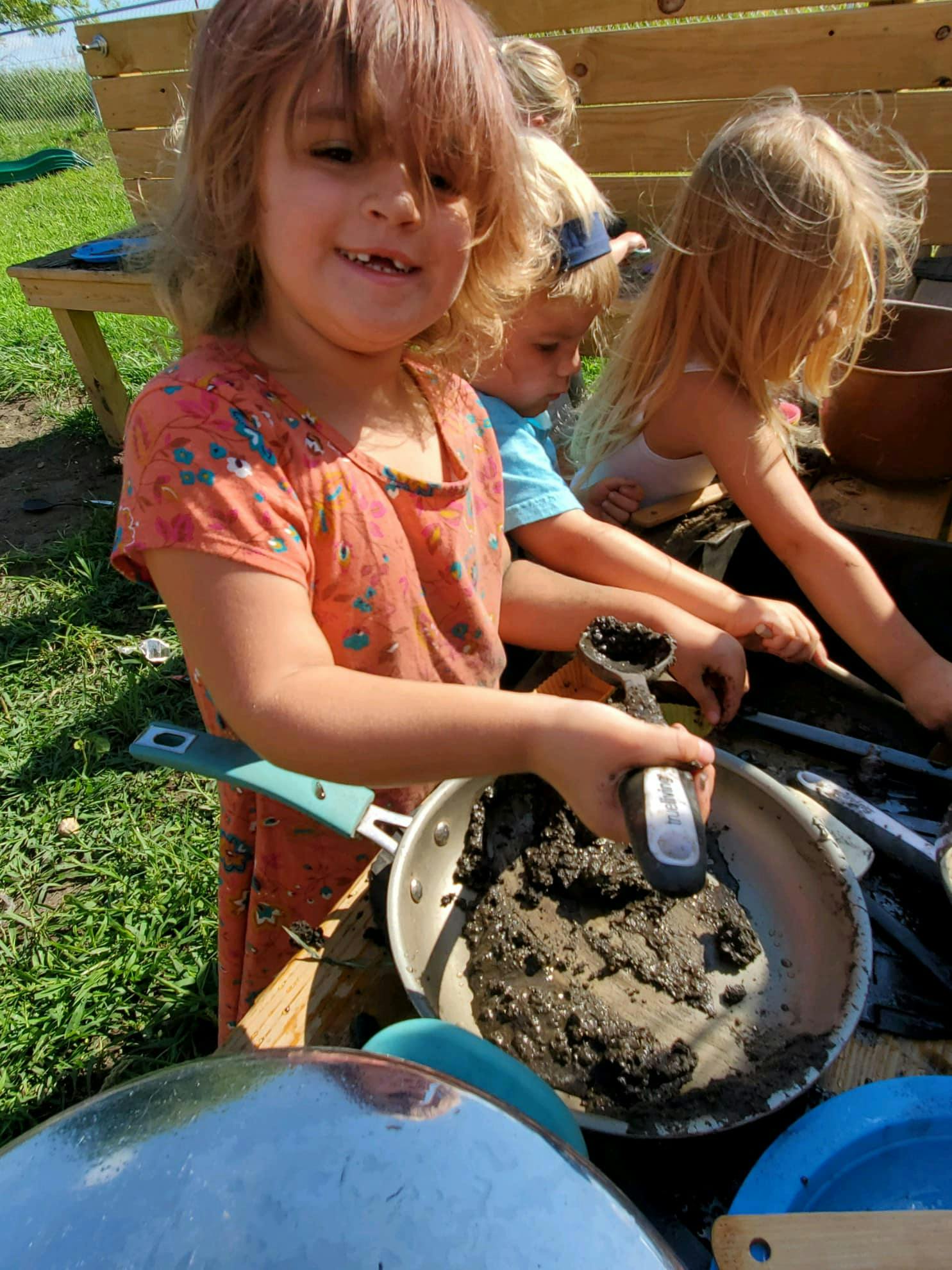 Child holding a spatula in a pan of mud with other children nearby