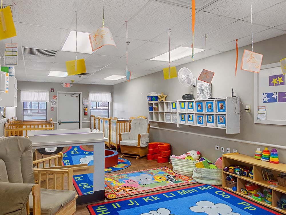 A childcare room with cribs, play mats, shelves of toys, and hanging decorations