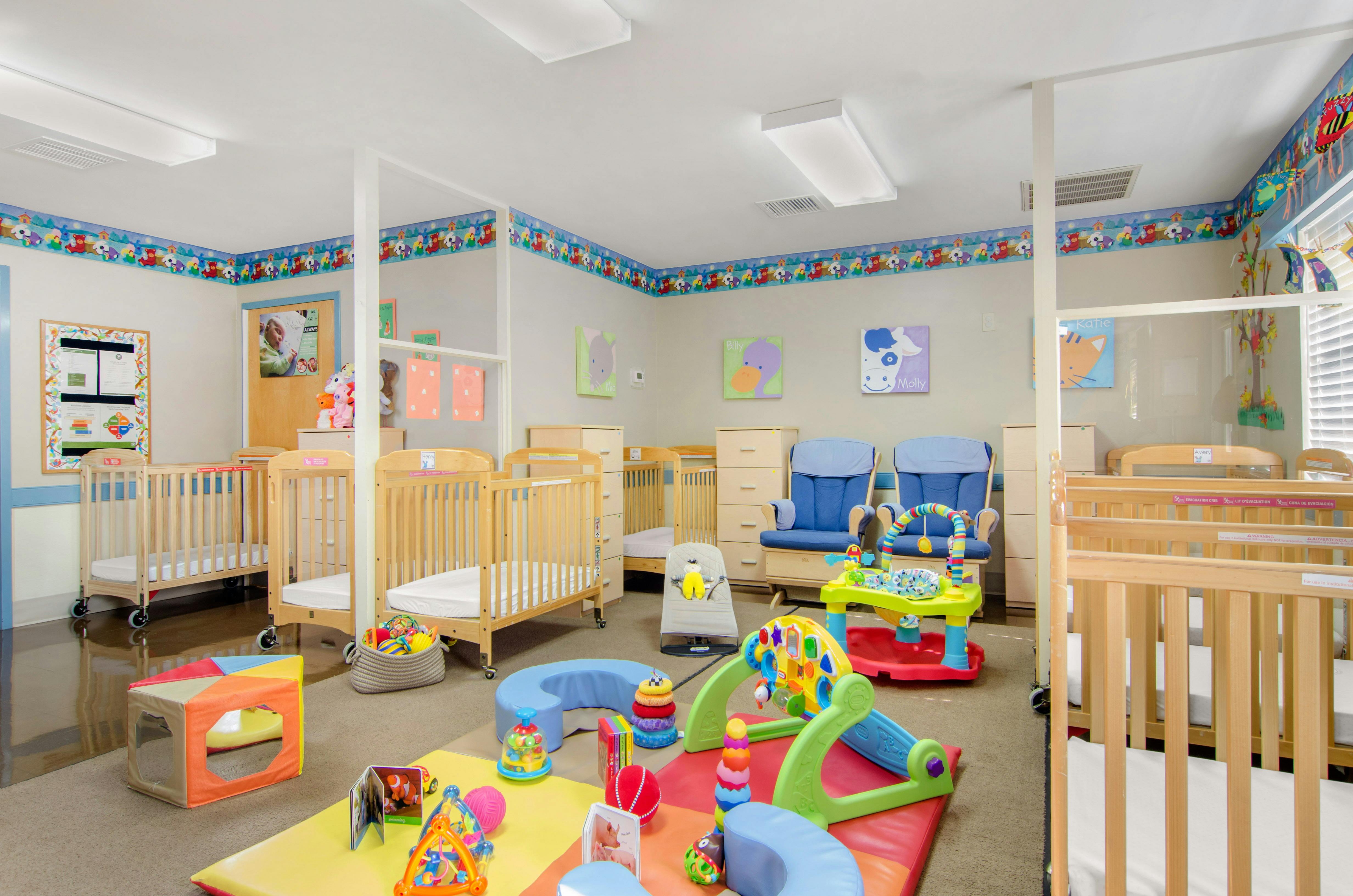 A colorful childcare room with several cribs, two blue rocking chairs, and toys