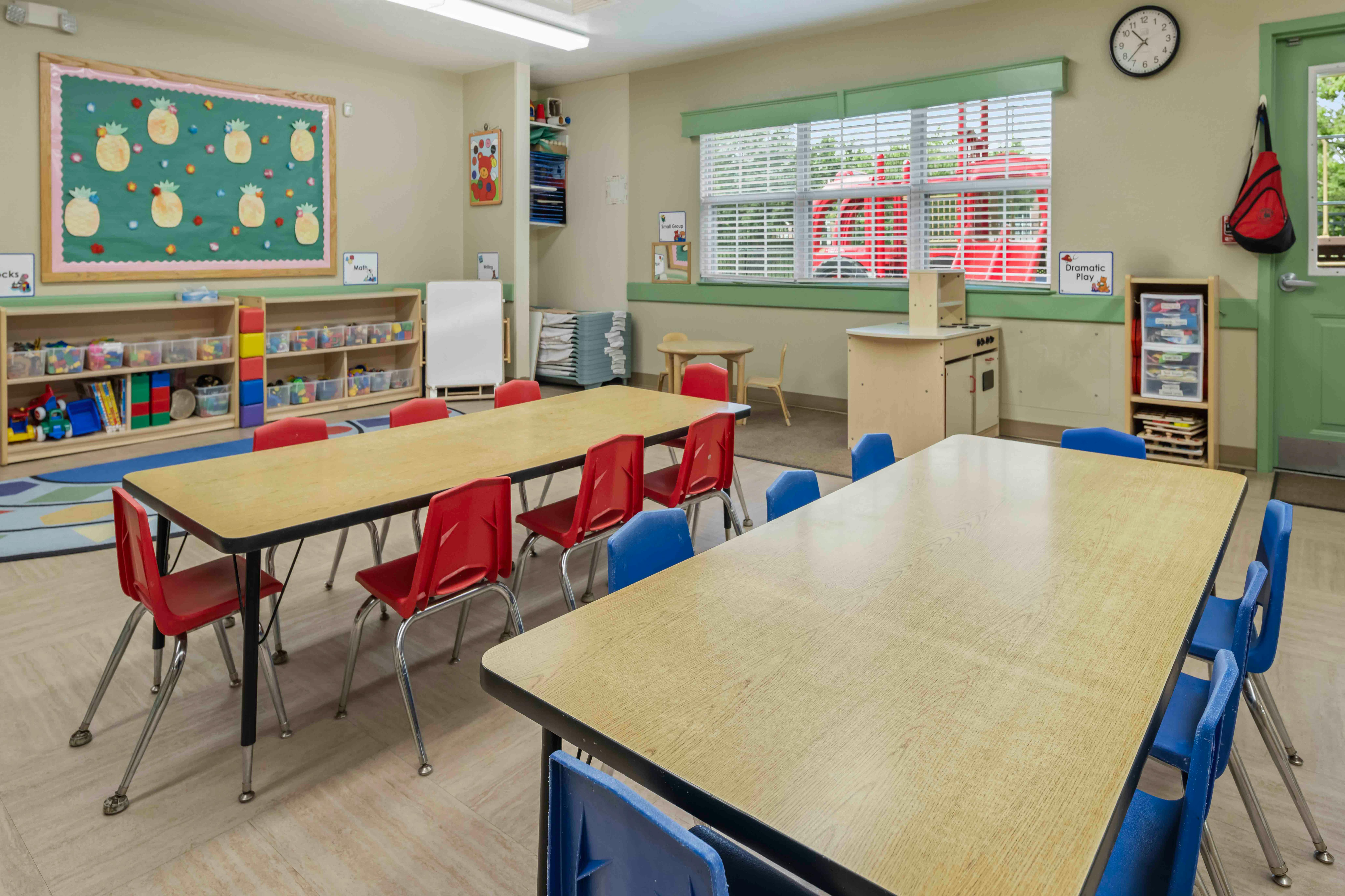 Classroom with multiple long tables and chairs, shelves with toys and books