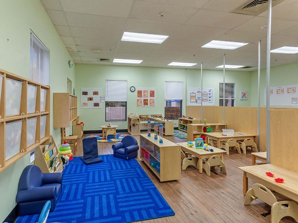 Classroom with small tables, chairs, shelving units, and a blue rug