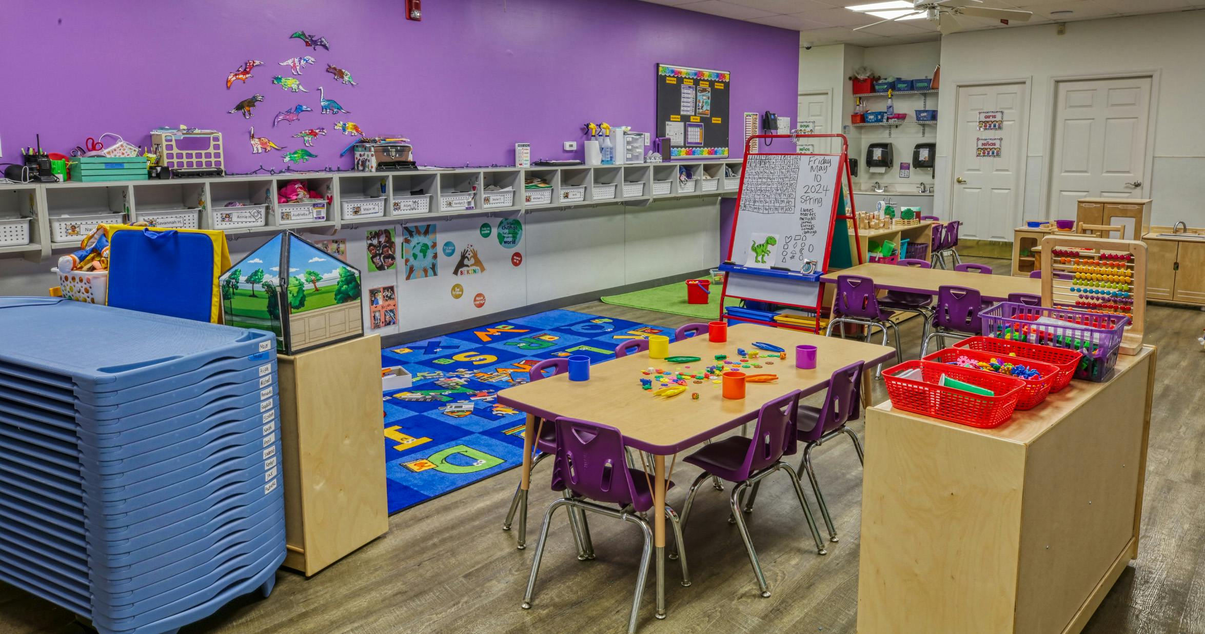Classroom with small tables and chairs, a play rug, and shelves with cubbies